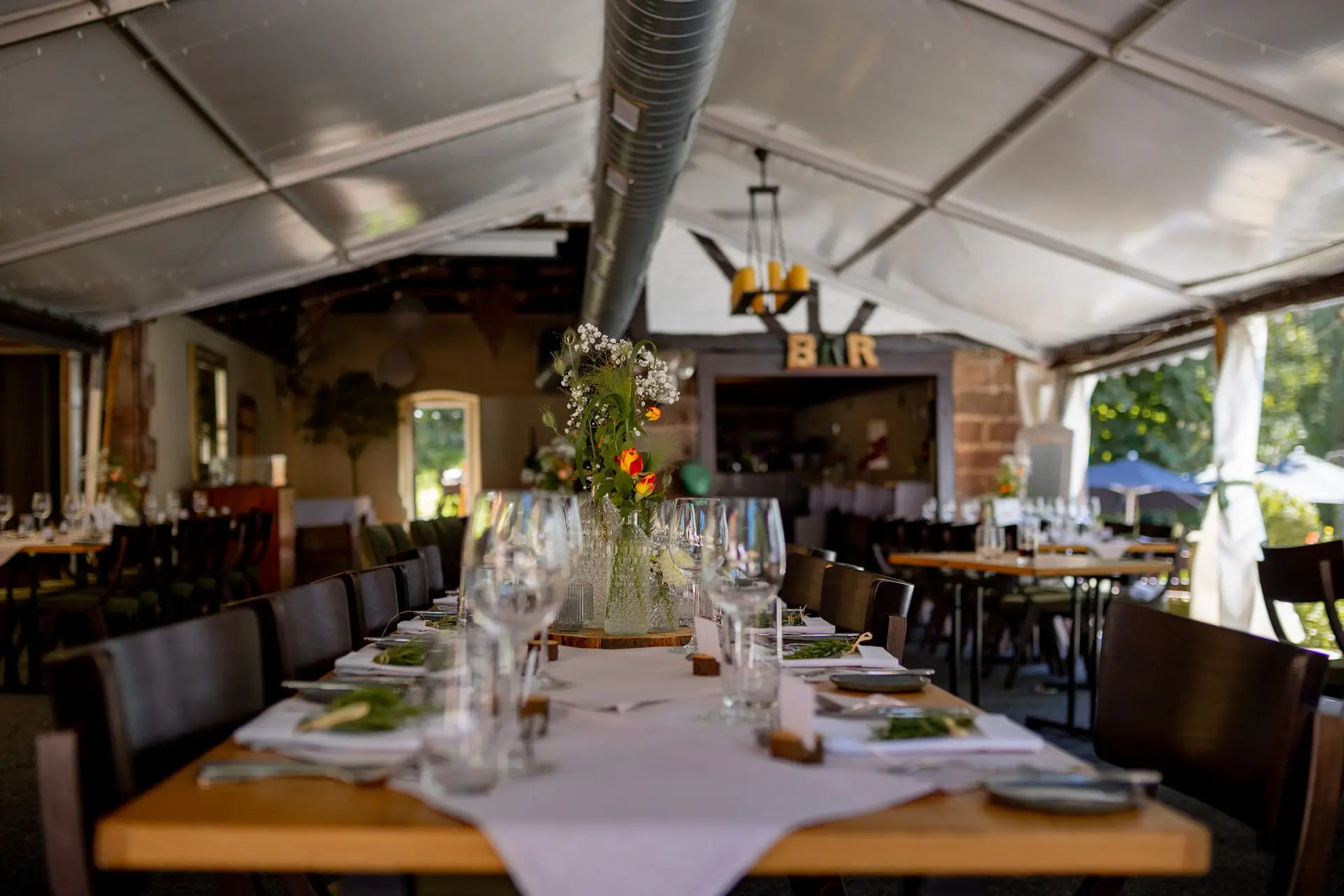 Langer Holztisch im hellen Zelt mit weißer Blumenvase und Bar-Schild der Location
