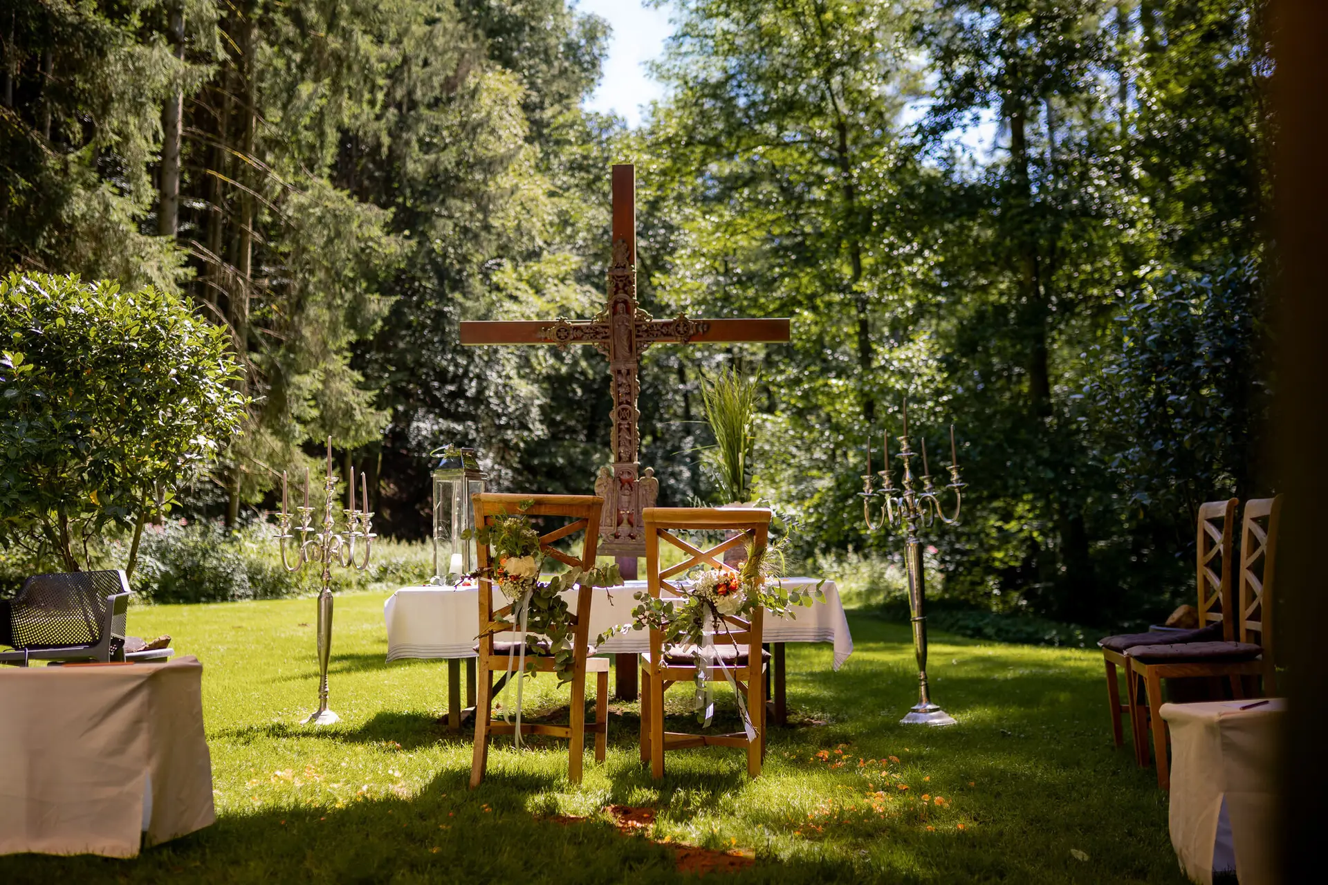 Romantische Trauungskulisse im Wald – Holzkreuz, floraler Schmuck, Stühle und Kerzenleuchter unter freiem Himmel