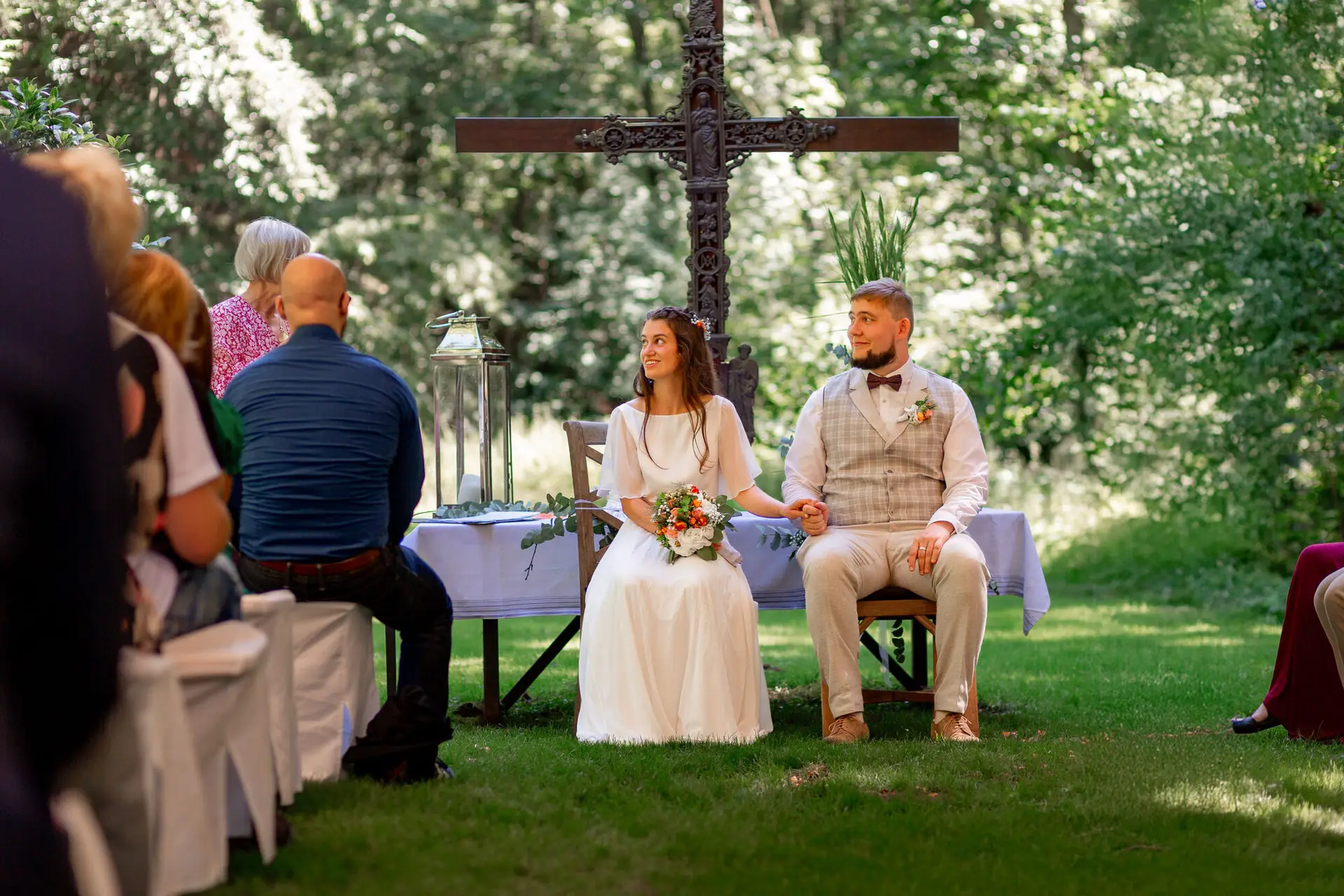 Brautpaar sitzt gemeinsam bei der Zeremonie vor dem Holzkreuz, Gäste umgeben sie im Halbkreis