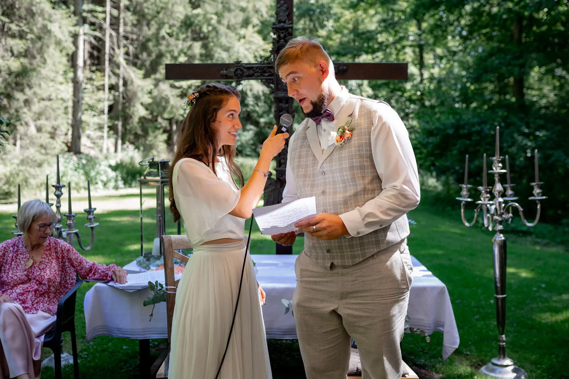 Bräutigam spricht seine Gelübde mit Mikrofon, Braut reicht ihm lächelnd den Zettel
