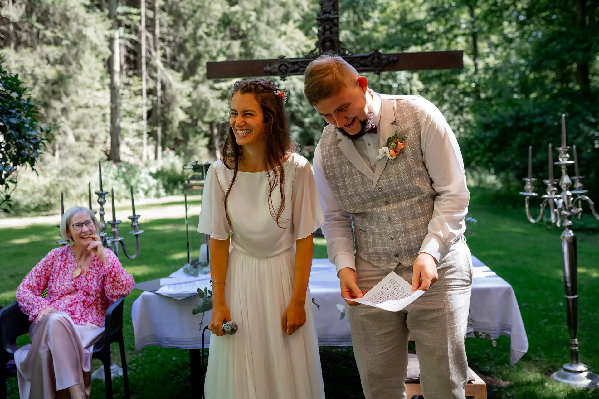 Brautpaar steht gemeinsam vor dem Holzkreuz auf der Wiese, Traurednerin lächelt im Hintergrund