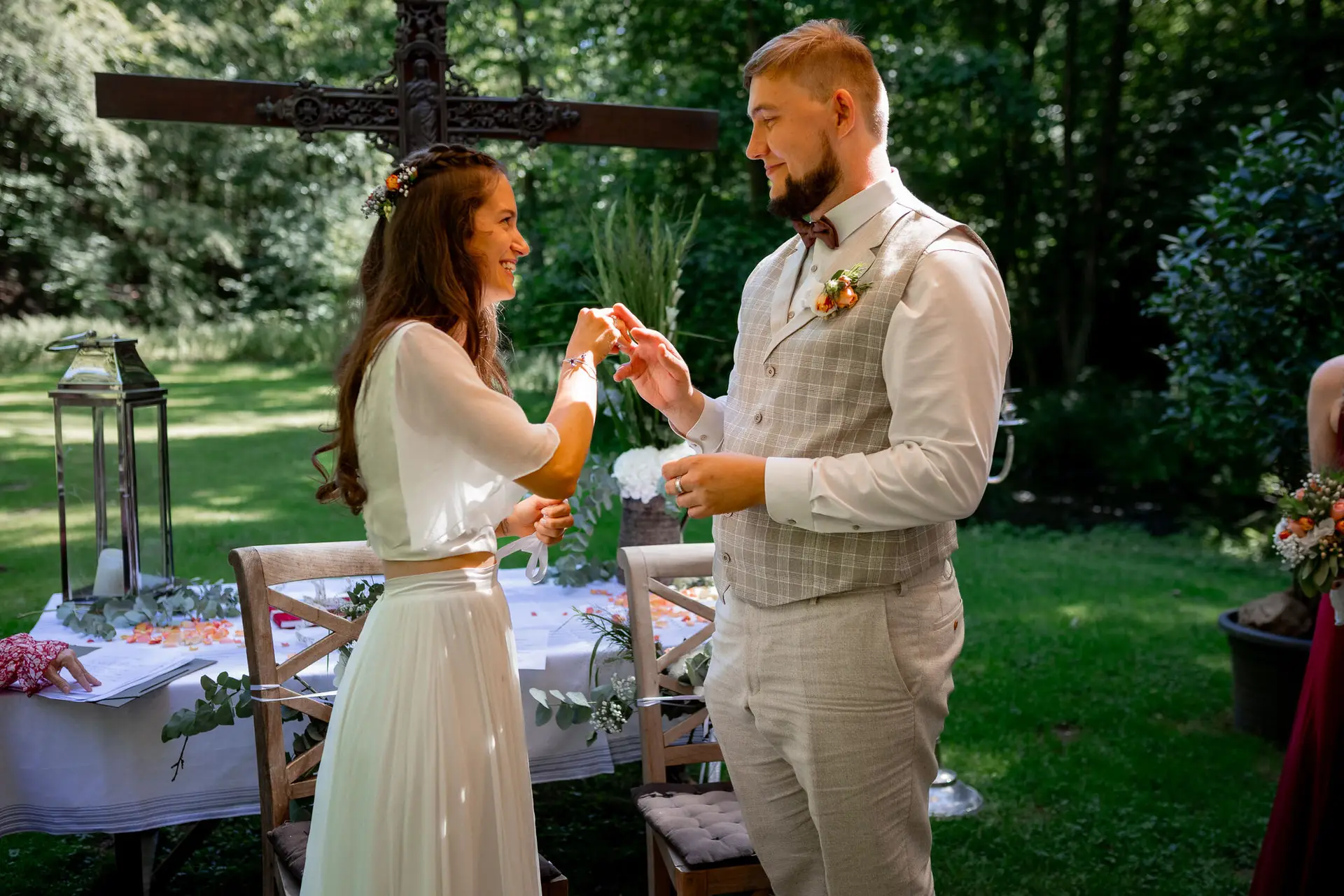 Brautpaar reicht sich gegenseitig eine Kette vor dem Holzkreuz – persönliches Zeremonieritual der beiden