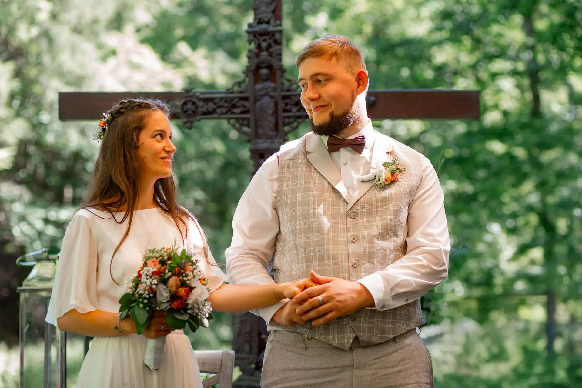 Brautpaar blickt sich zärtlich an vor dem verzierten Metallkreuz – stiller Moment nach der Trauung