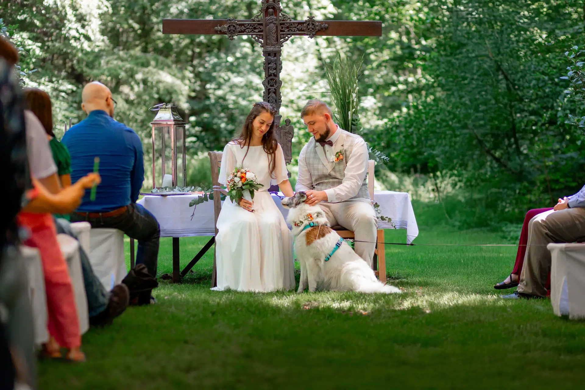 Braut und Bräutigam sitzen auf Bank mit der weißen Hündin, Gäste gratulieren im Hintergrund