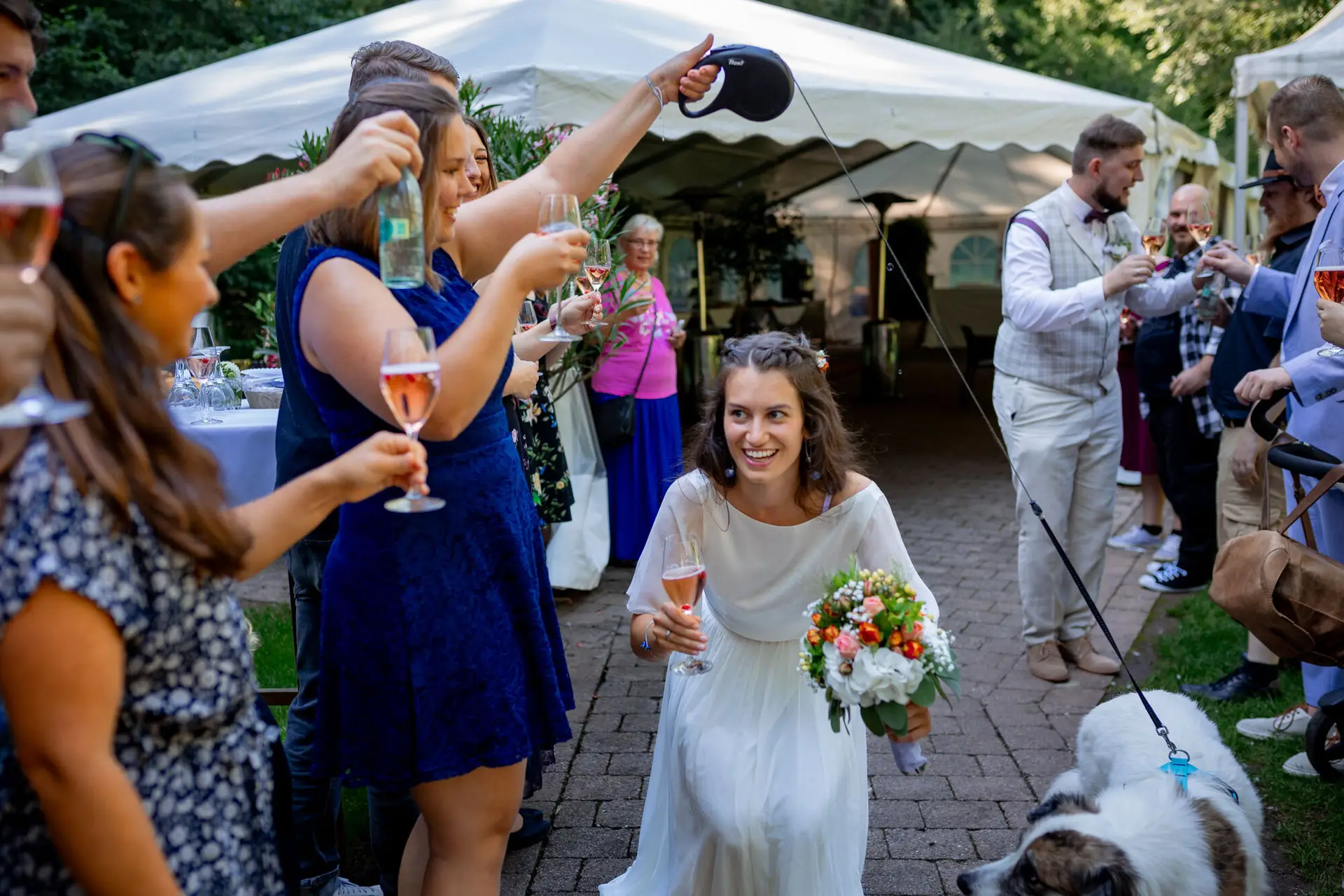 Braut lacht herzlich mit Gästen beim Champagner-Empfang im Freien der Heidersbacher Mühle