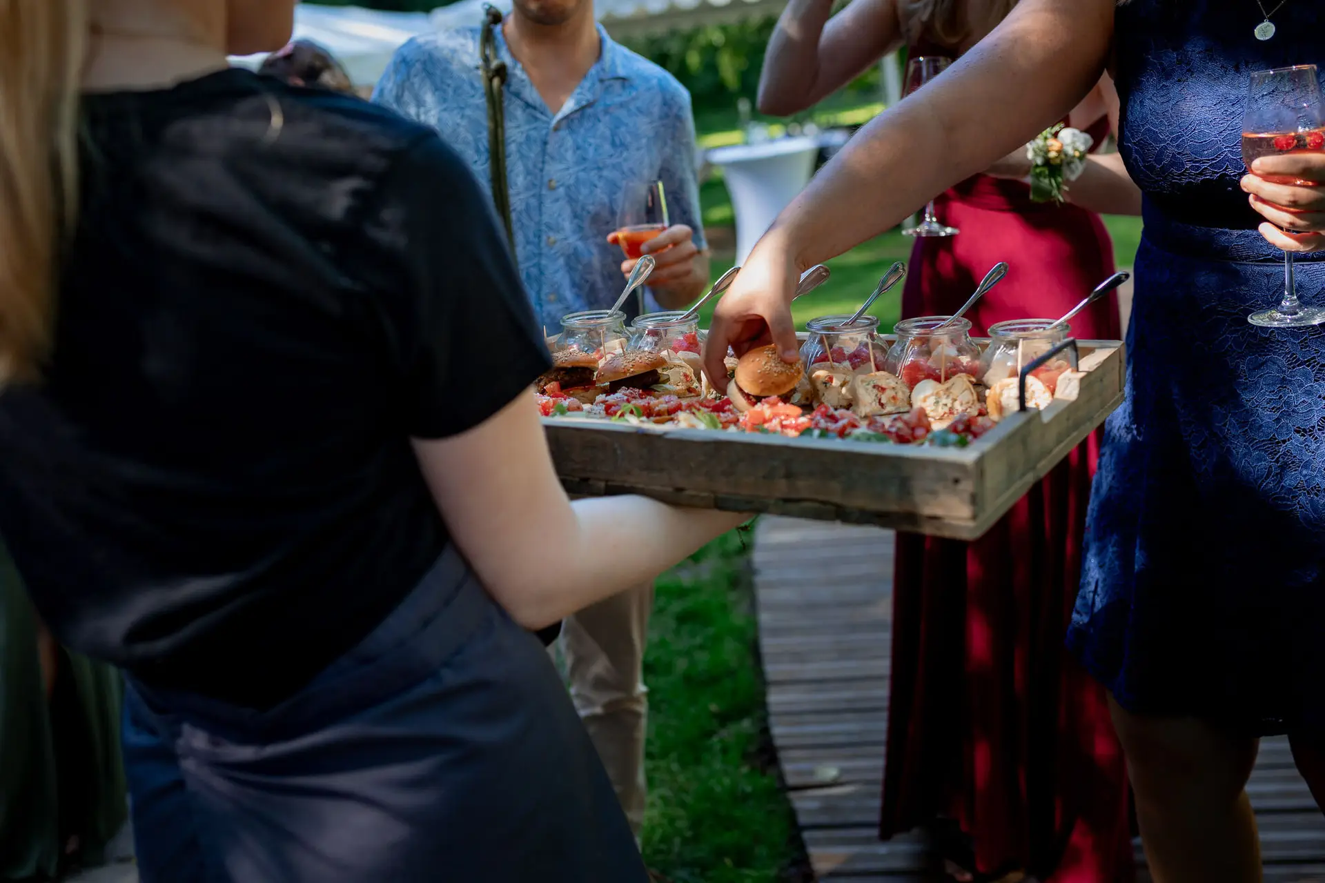 Kellner trägt rustikale Holzplatte mit Vorspeisen und Wein über den sonnigen Rasen