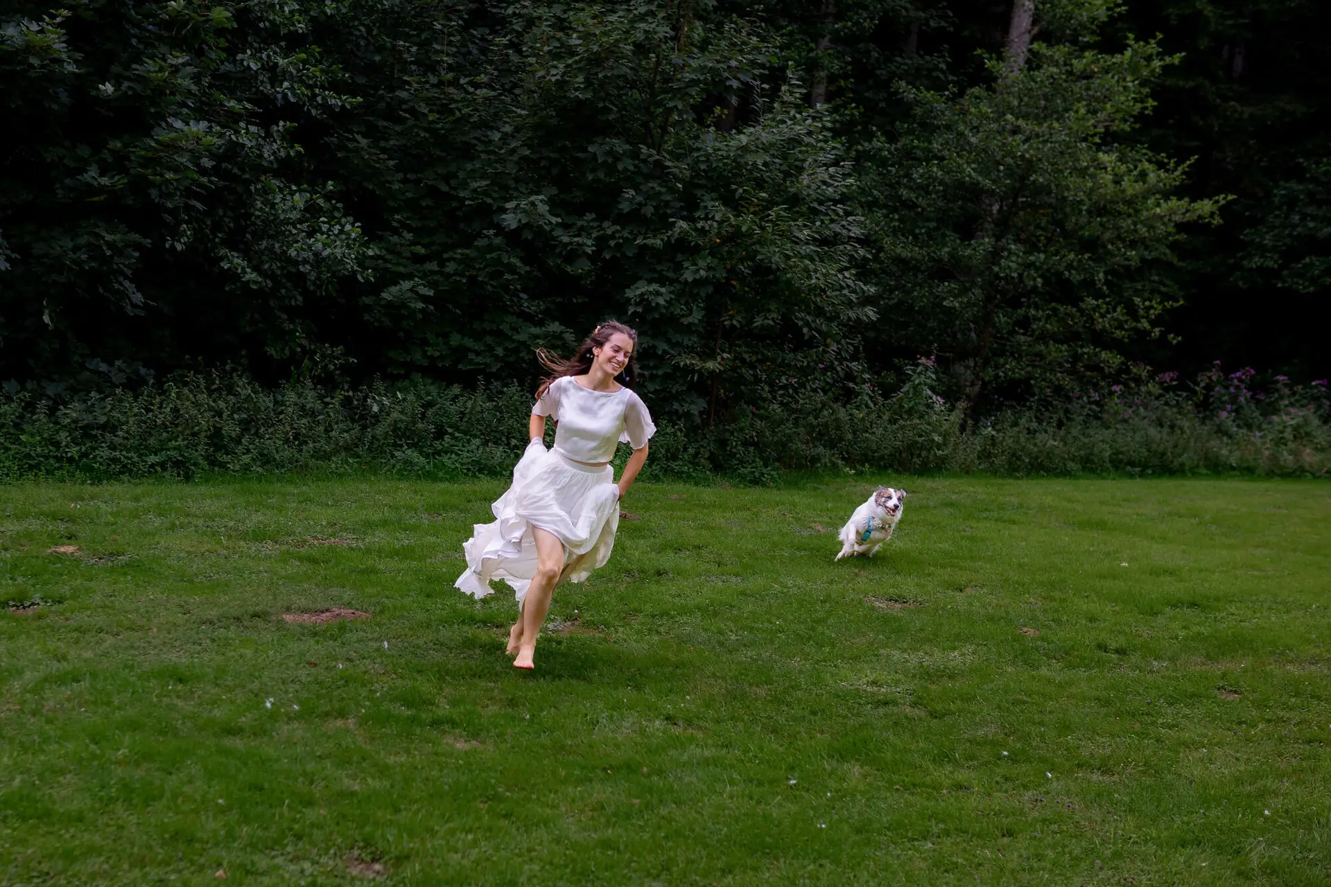 Braut rennt fröhlich durch die grüne Wiese, Hund folgt begeistert, Elztal-Wald im Hintergrund