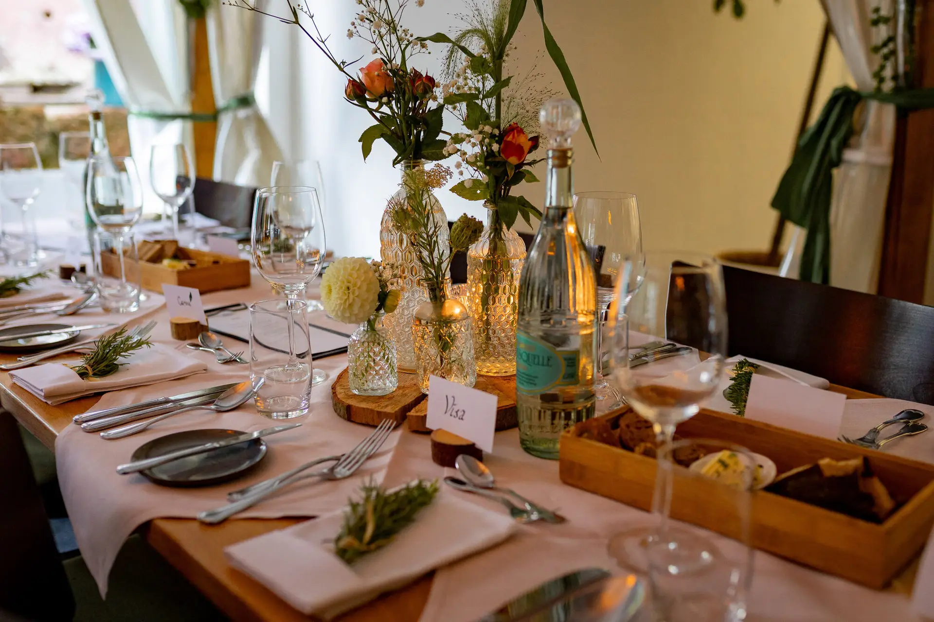 Langer Festtisch mit hellrosa Tischdecken und Holzscheiben, Gäste versammeln sich zum Abendessen