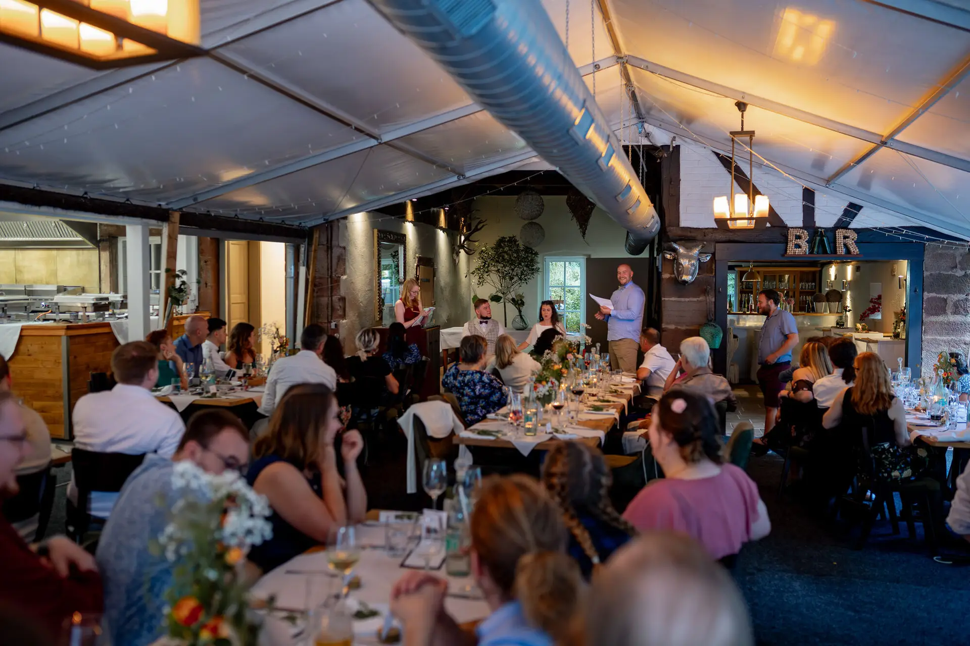 Große Hochzeitsgesellschaft beim Abendessen, Redner mit Mikrofon hält Ansprache an den Tischen