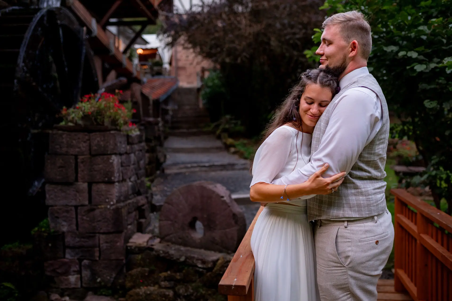 Brautpaar umarmt sich zärtlich beim alten Wasserrad im abendlichen Mühlengarten