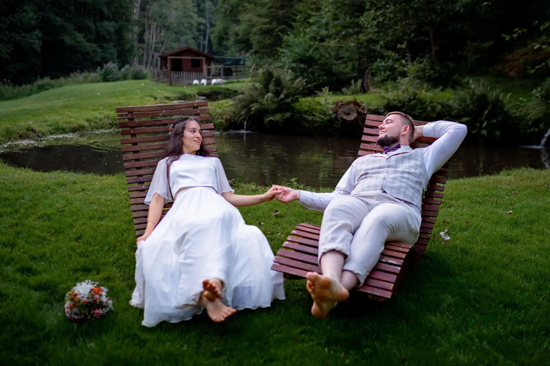 Brautpaar liegt auf Holzliegen am ruhigen Teich und hält Hände, Bäume und Natur ringsum