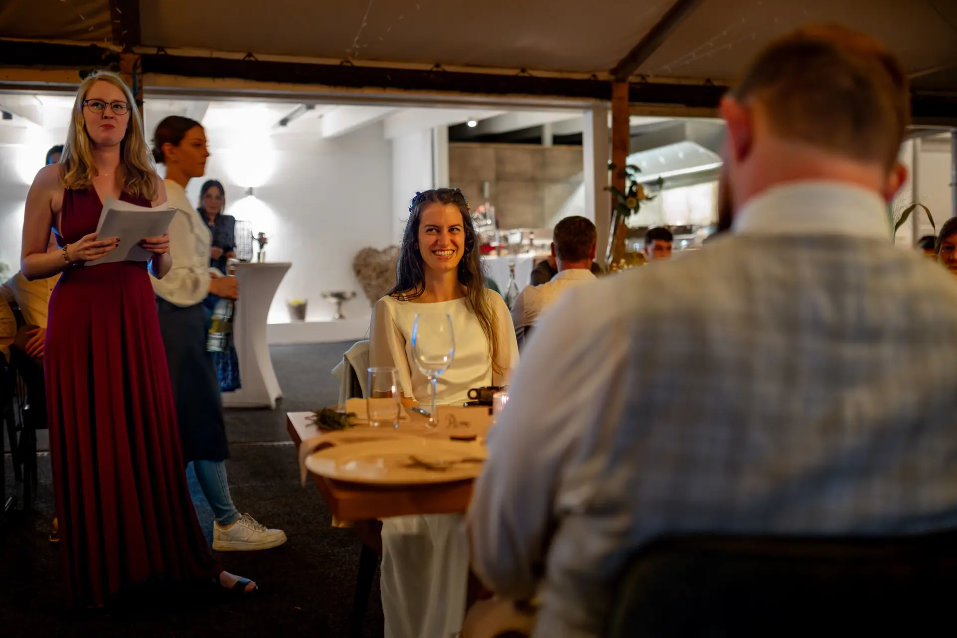 Braut lächelt glücklich in die Kamera beim Hochzeitsabend, Bräutigam schaut aus dem Hintergrund