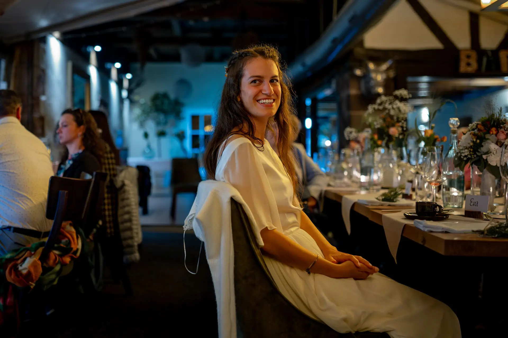 Braut sitzt entspannt am Abendtisch und genießt den Moment, Gäste feiern im Hintergrund