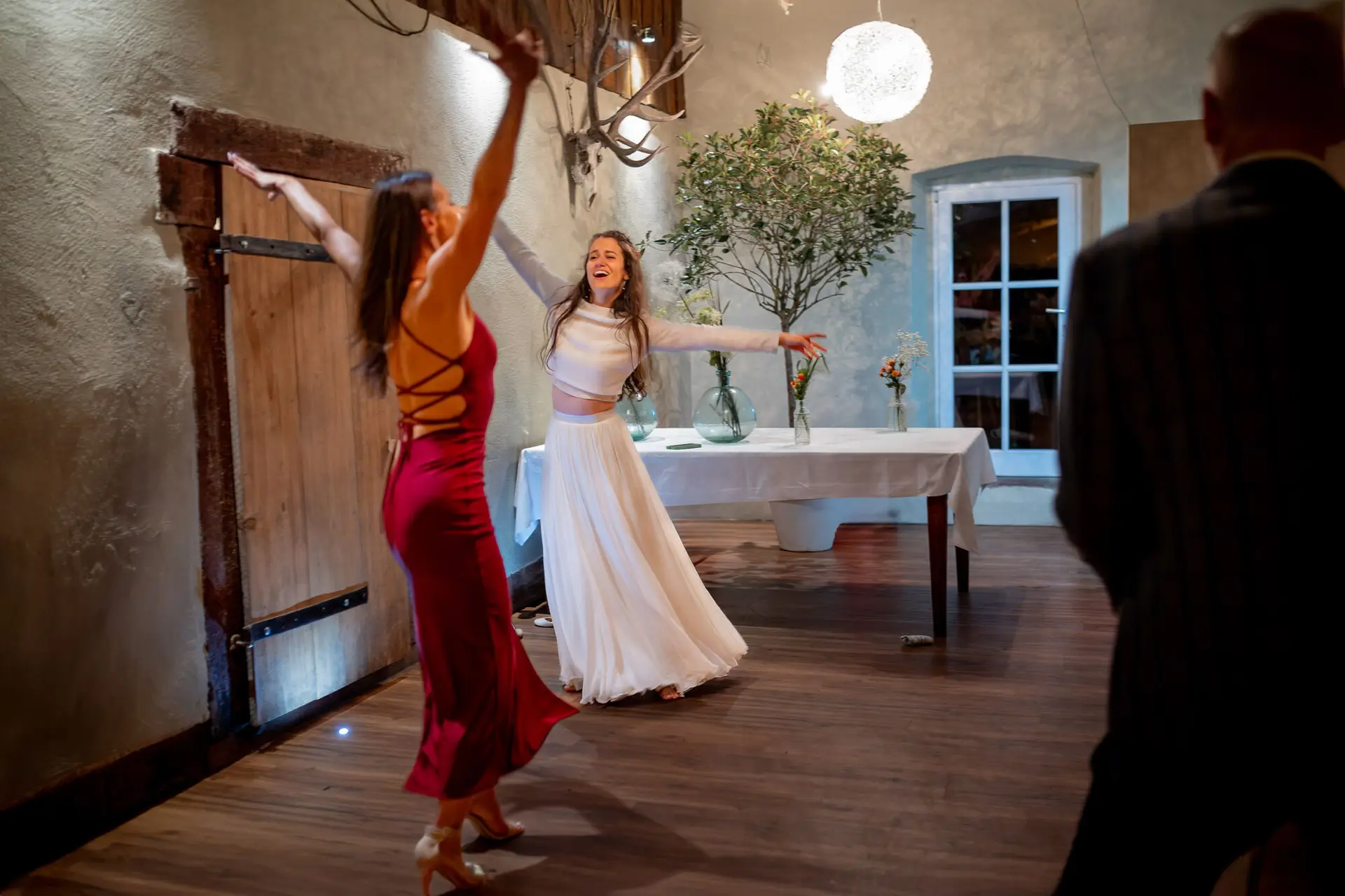 Braut springt freudig in die Luft, Brautjungfer in Rot im Hintergrund – Energie pur bis in die Nacht