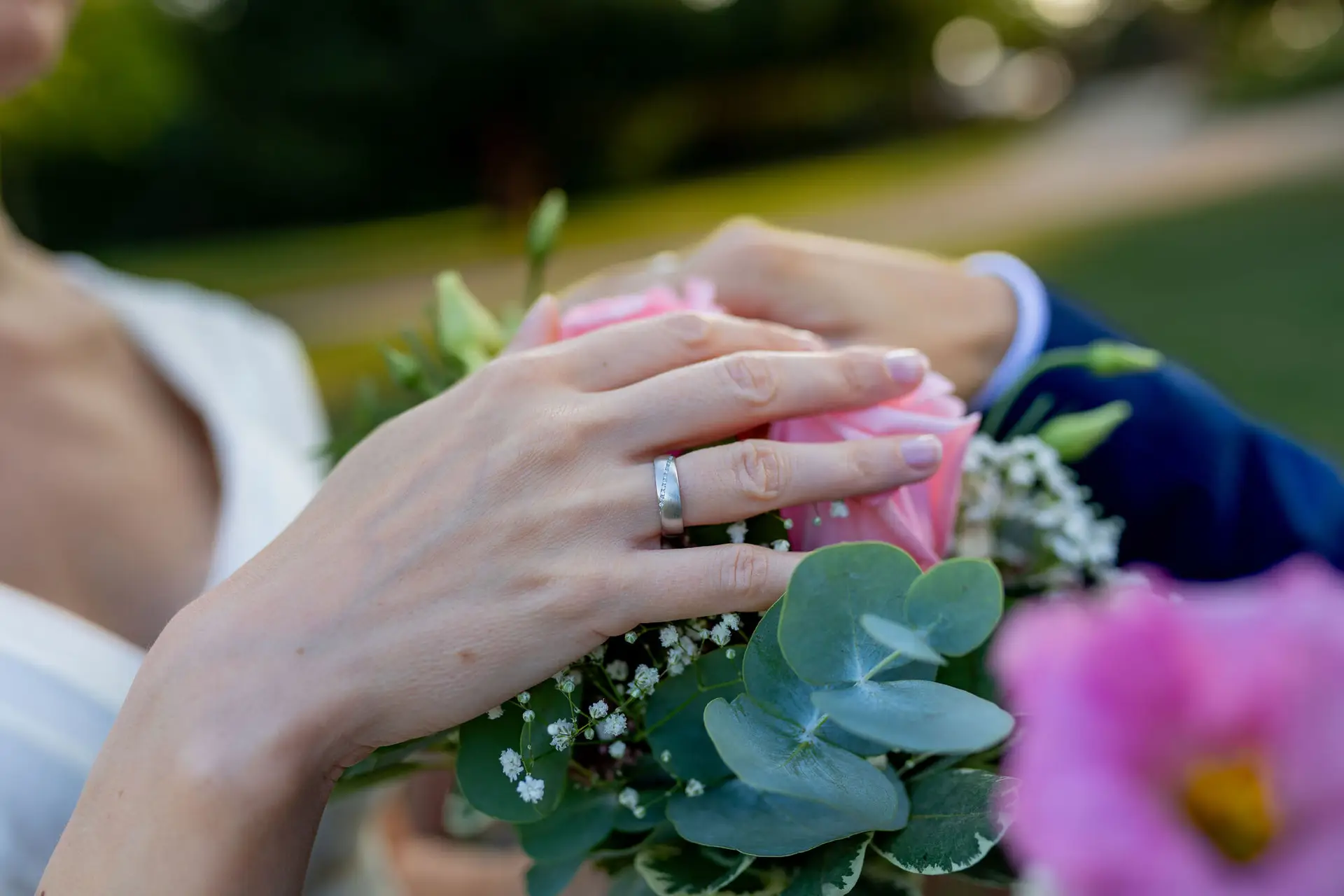 Braut hält Strauß aus rosa Rosen und Eukalyptus, Brautpaar hält sich an den Händen im Garten