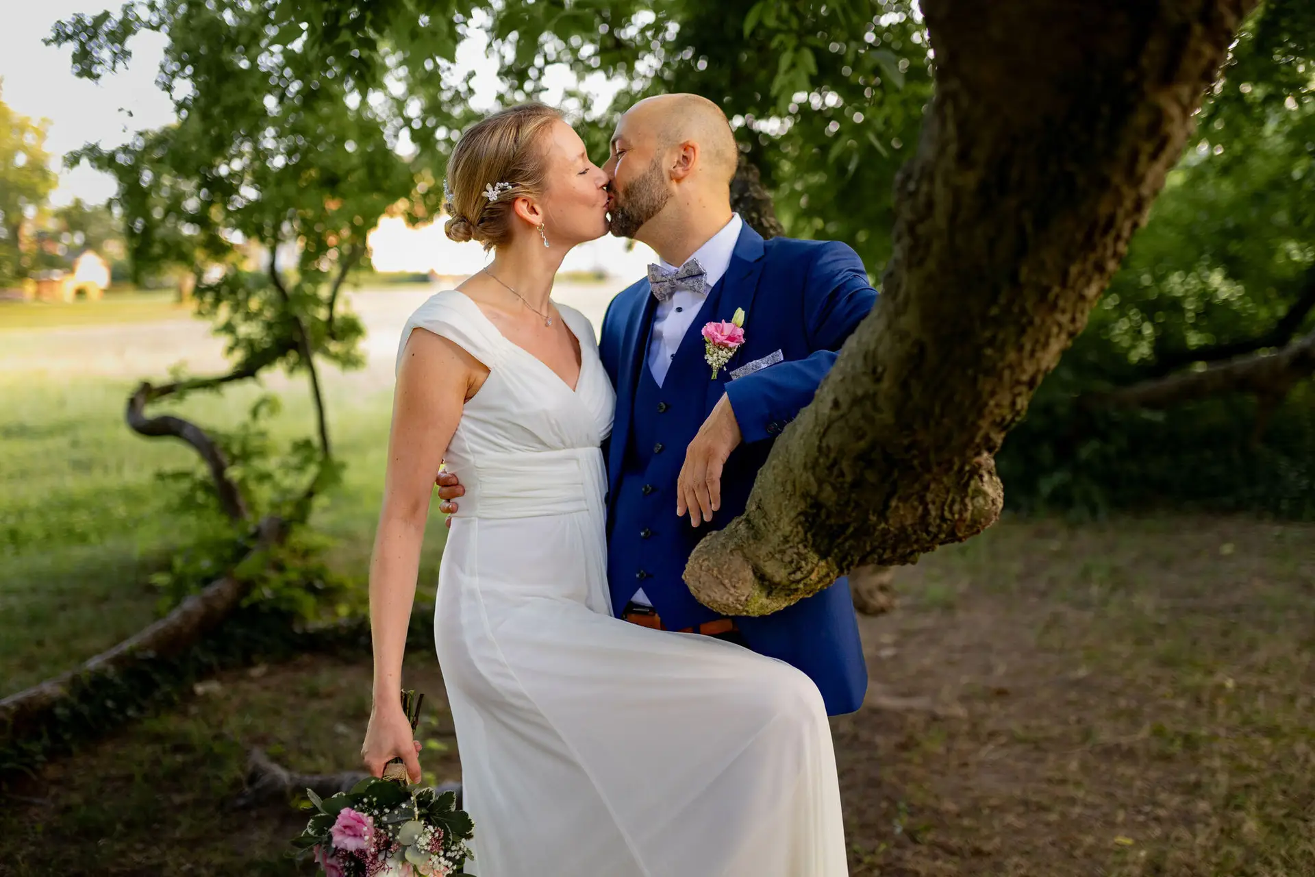 Brautpaar küsst sich unter den Ästen eines alten Baumes auf grüner Wiese im Schlossgarten Herrnsheim