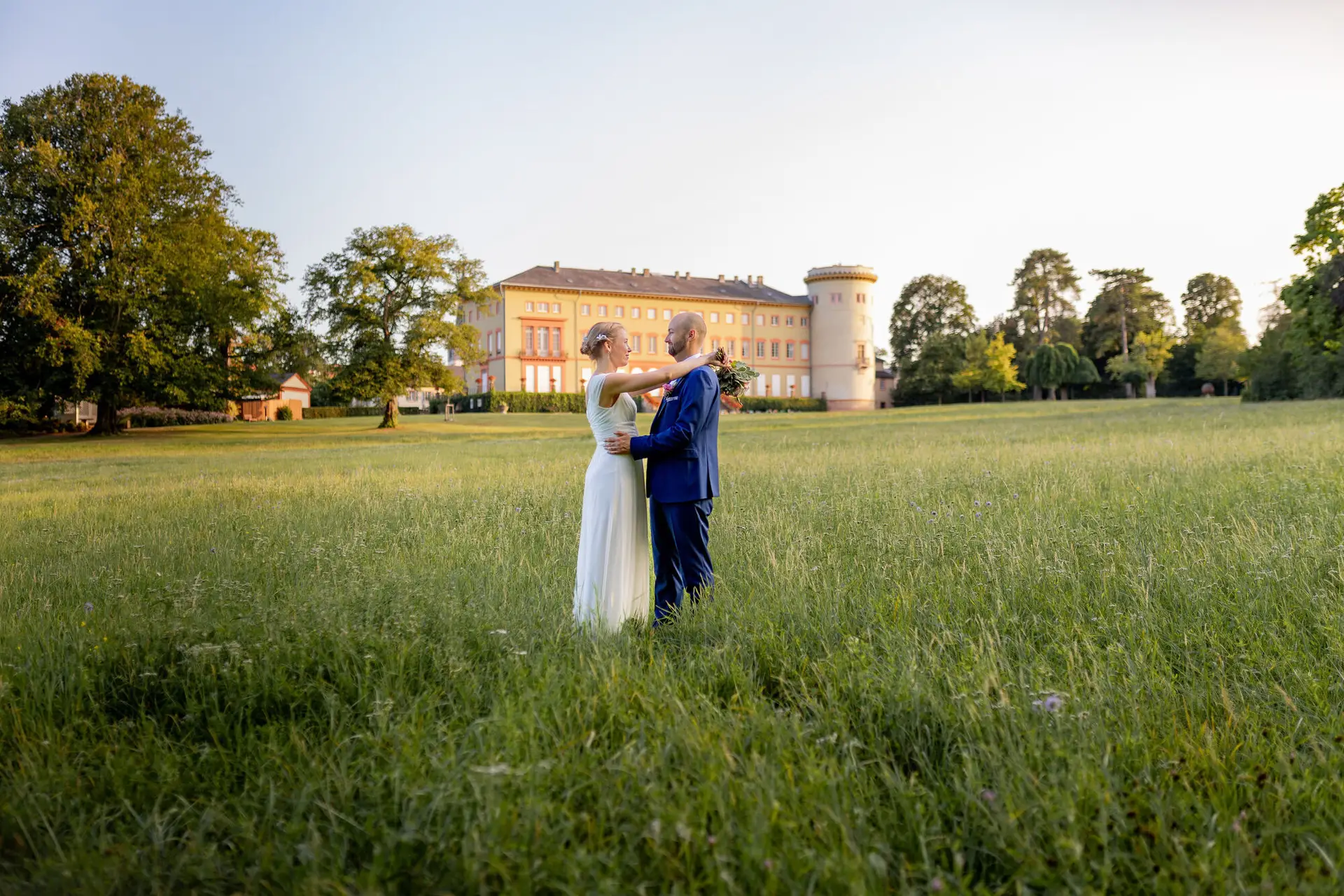 Brautpaar umarmt sich eng in hohem grünen Gras, im Hintergrund das gelbe Schloss Herrnsheim