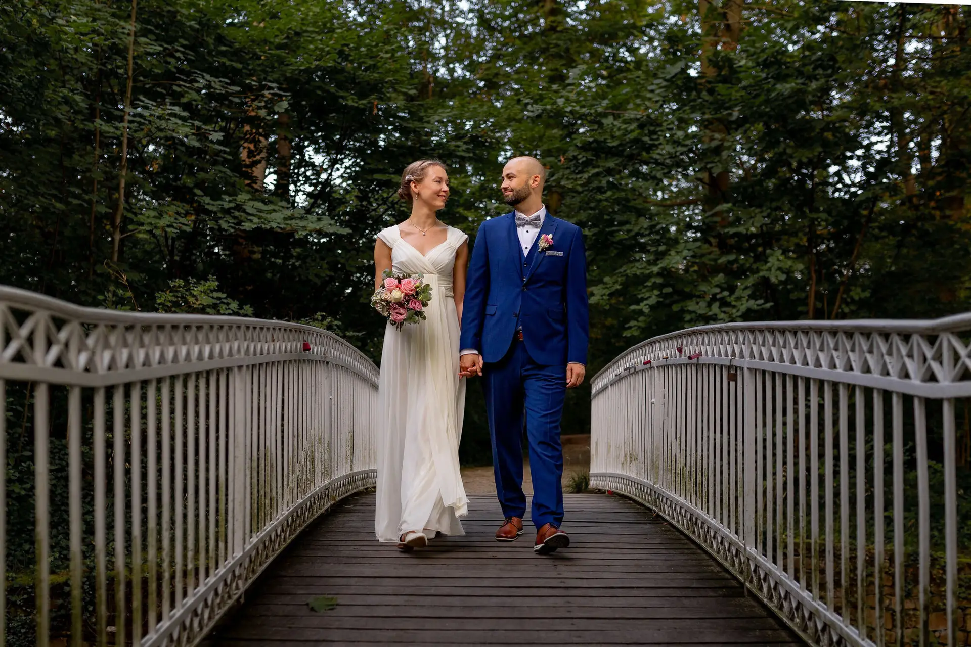 Brautpaar geht Hand in Hand über eine Holzbrücke durch einen dichten Waldbereich