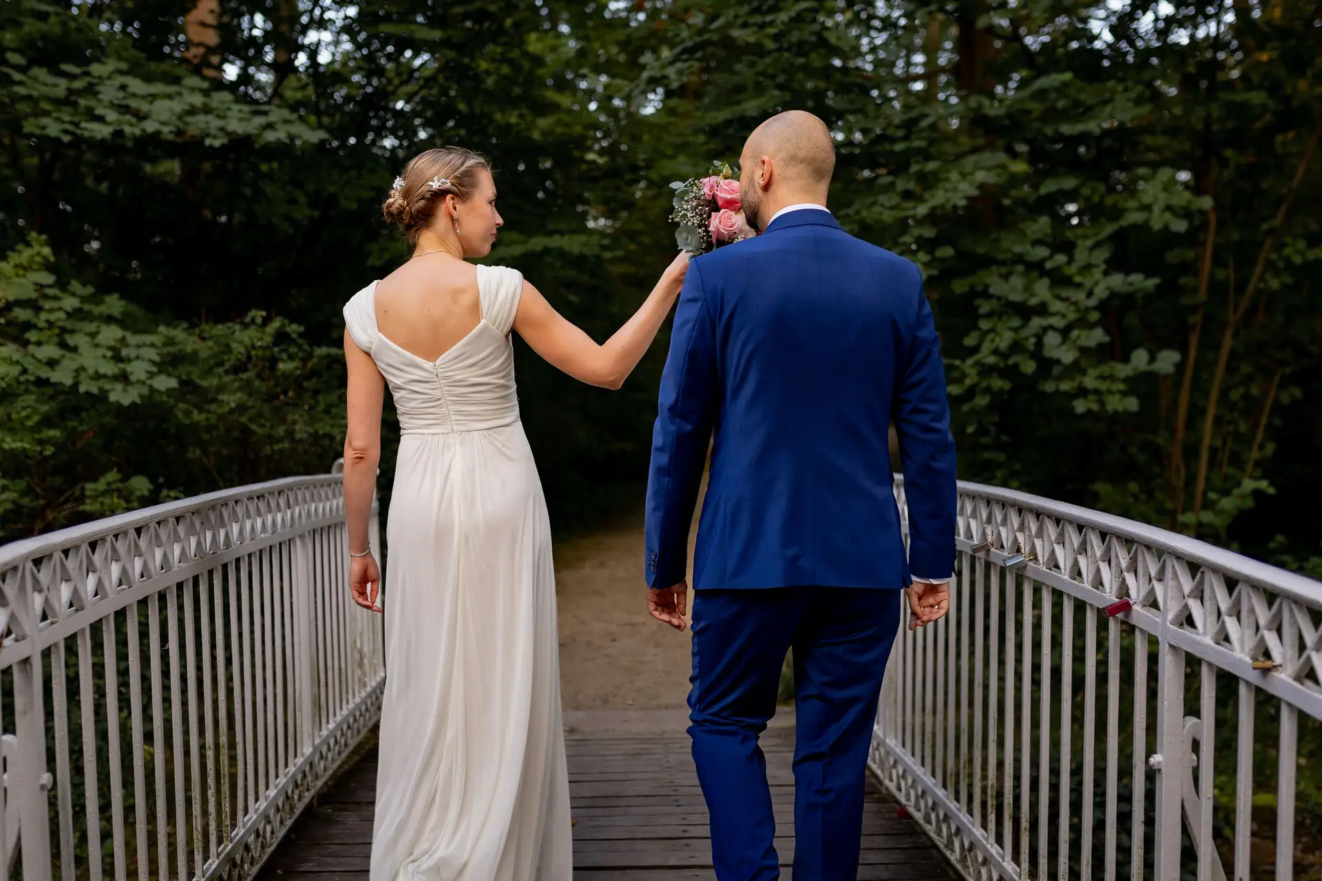 Brautpaar geht Hand in Hand über die Holzbrücke im Wald, Braut im weißen Kleid, Bräutigam im dunkelblauen Anzug