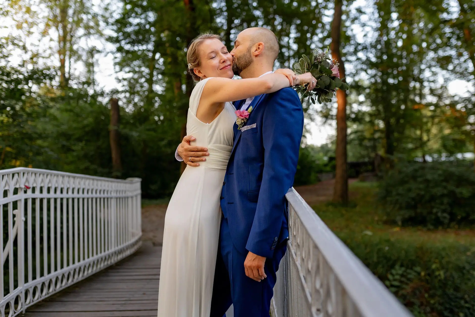 Bräutigam umarmt Braut auf der Holzbrücke im Wald, Braut hält kleinen Blumenstrauß