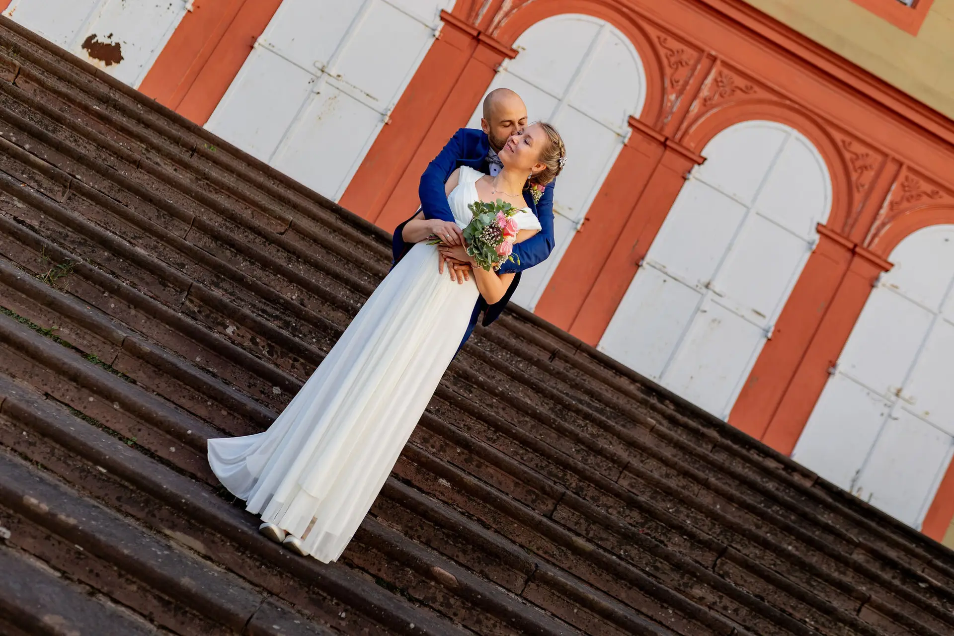 Brautpaar küsst sich auf den Stufen eines historischen Backsteingebäudes im Schlossgarten