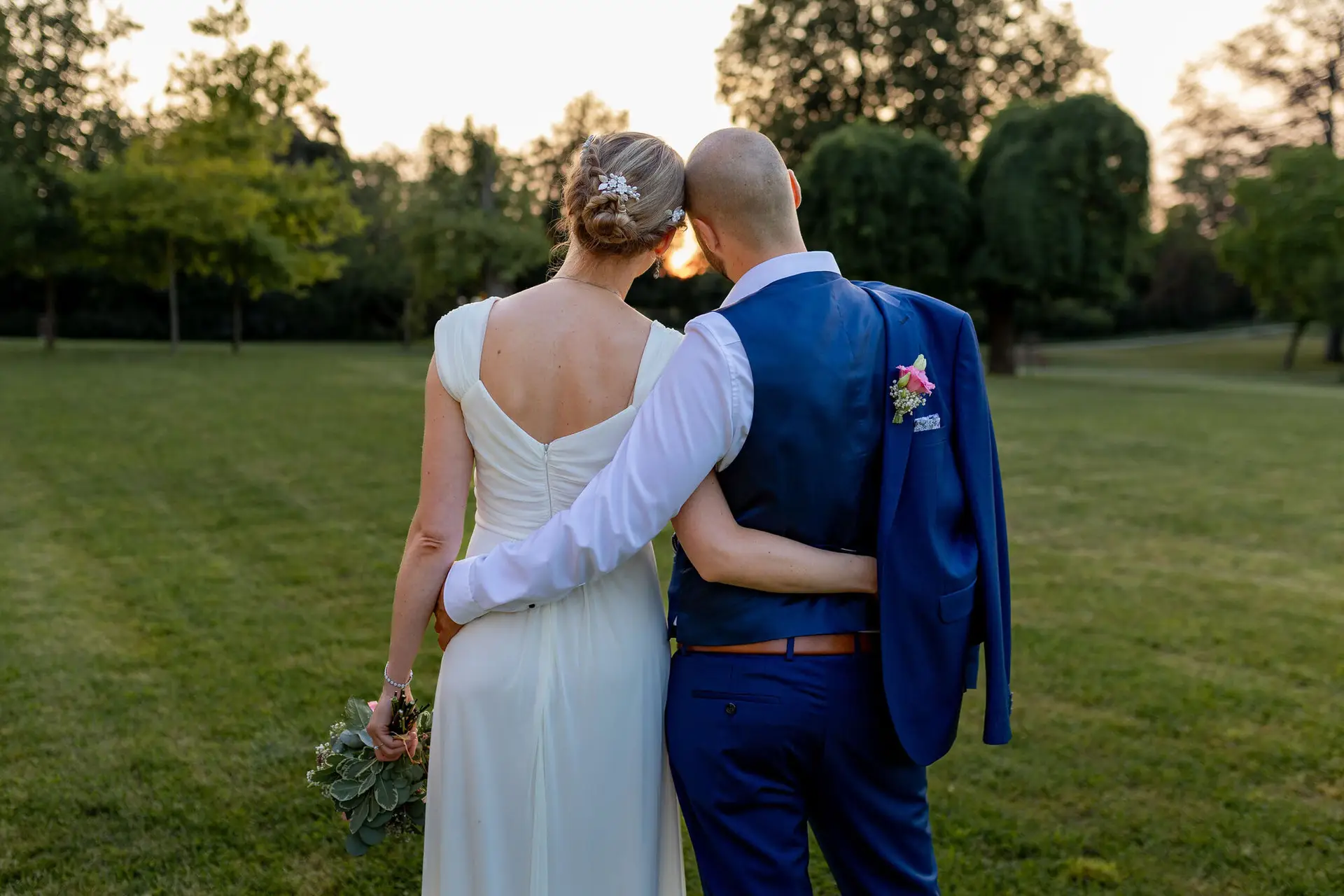 Brautpaar von hinten auf grüner Wiese zwischen Bäumen im Abendlicht des Schlossgartens Herrnsheim