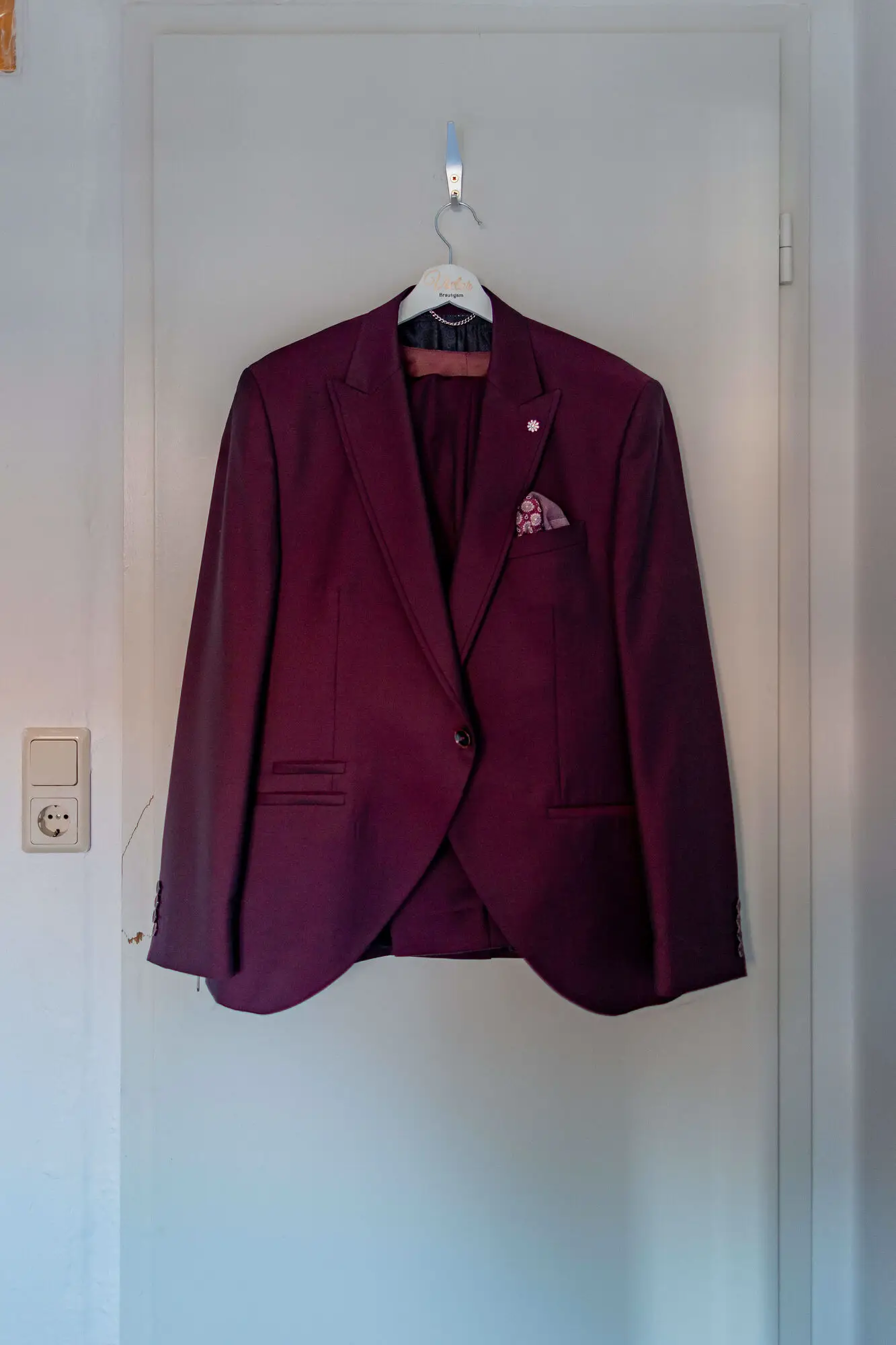 Bordeauxfarbenes Hochzeitssakko hängt an weißer Tür im Hotelzimmer