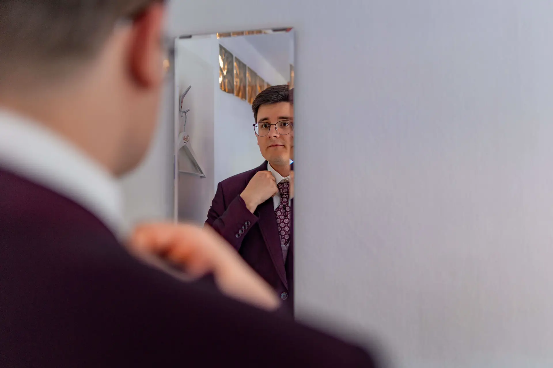 Bräutigam richtet Krawatte vor dem Spiegel im bordeaux Hochzeitsanzug