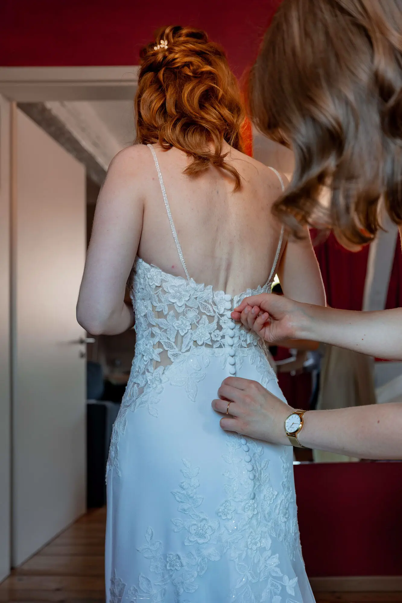 Braut wird von Trauzeugin in das Spitzenkleid mit offenen Rücken geholfen