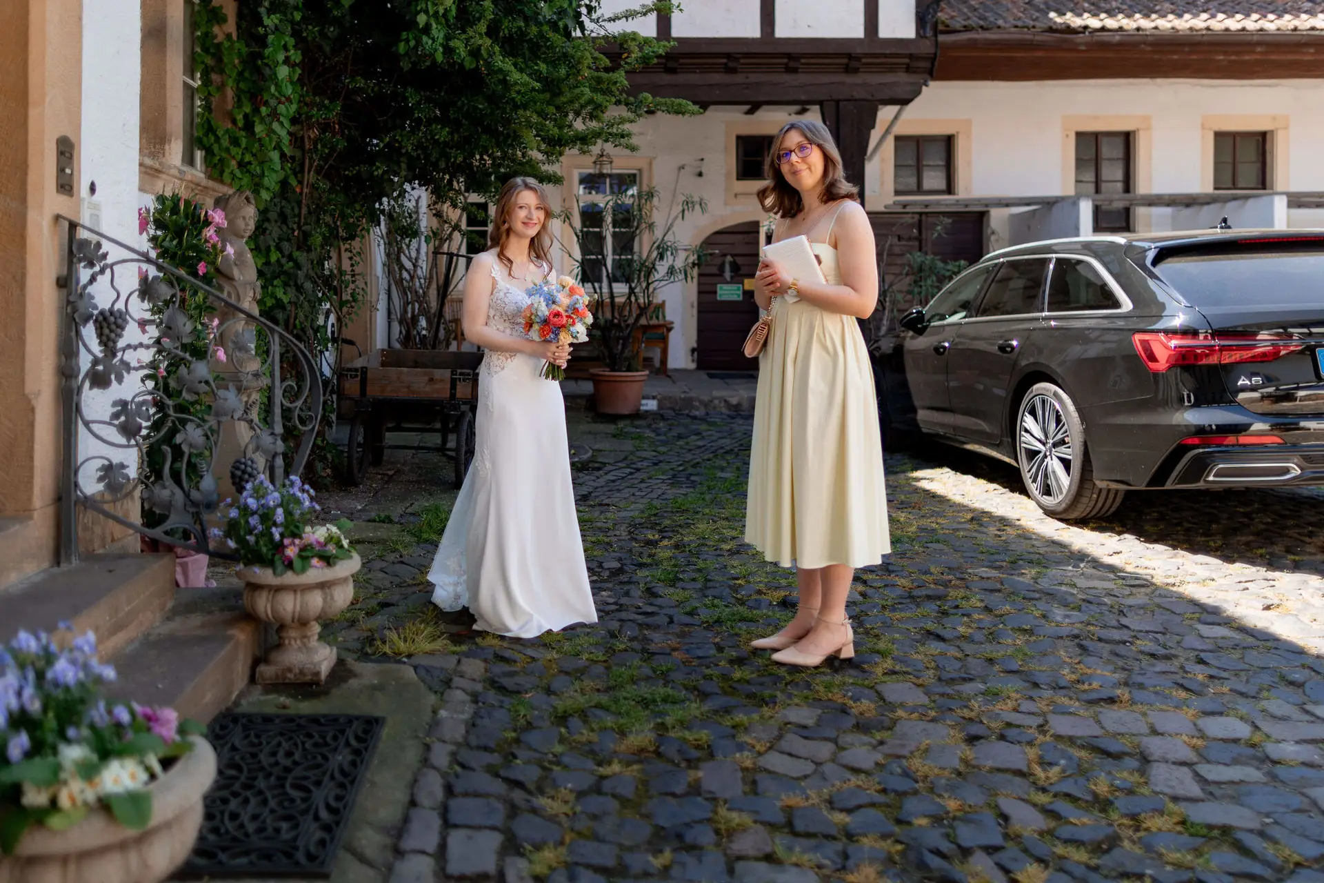 Braut mit buntem Brautstrauß und Trauzeugin in beigem Kleid im Innenhof des Weinguts