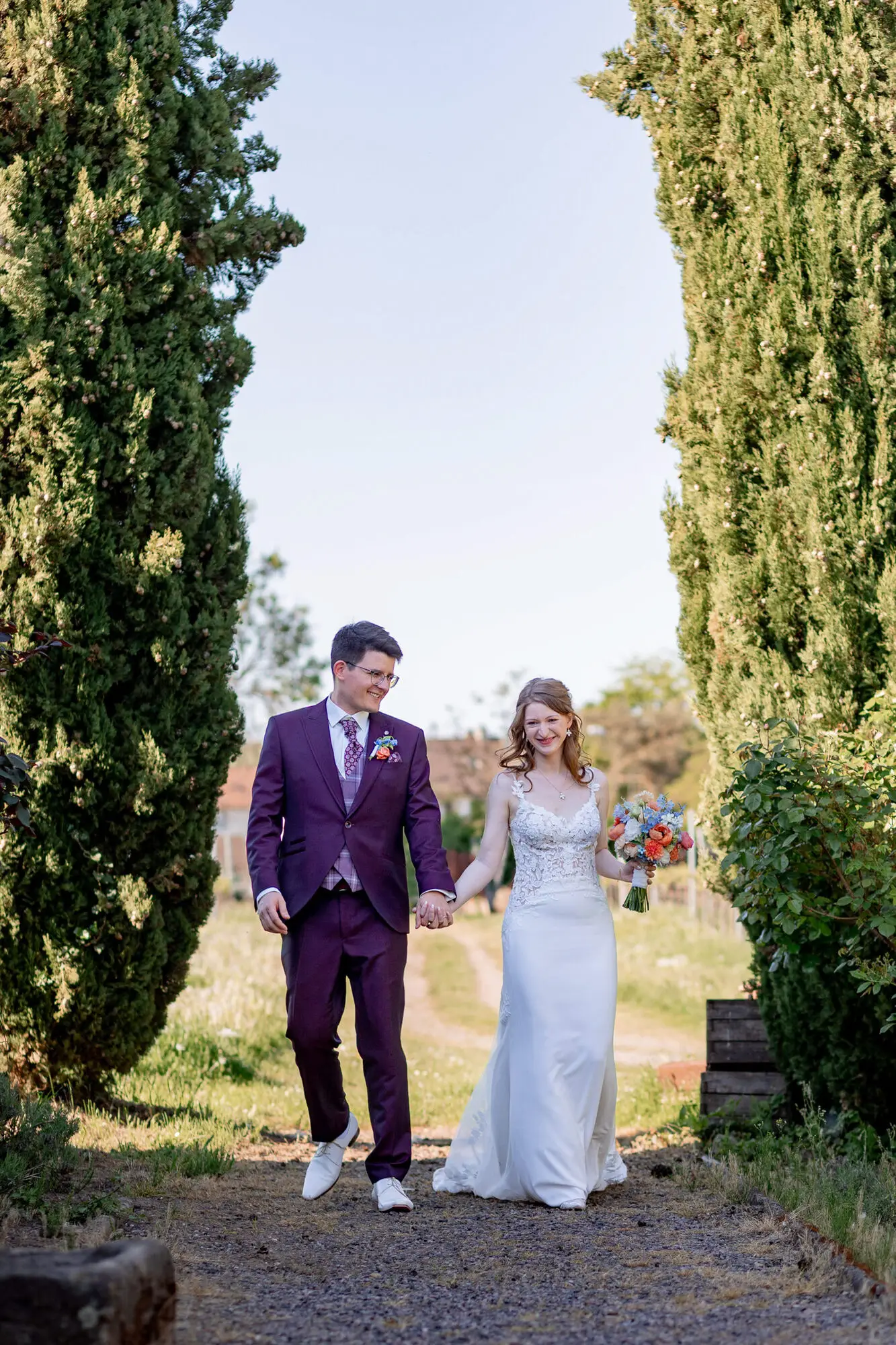 Brautpaar läuft Hand in Hand zwischen hohen Zypressen im Gutspark