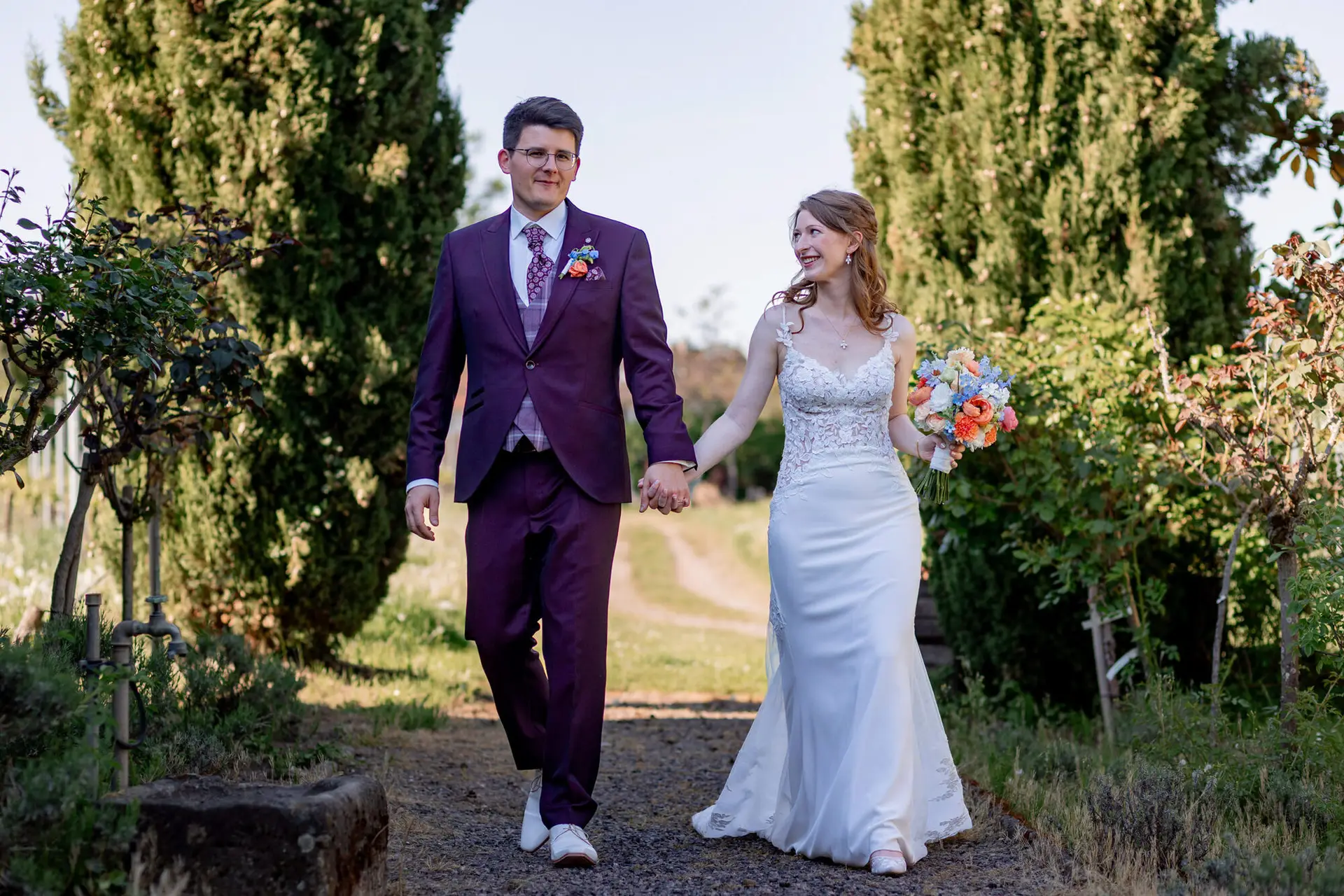 Brautpaar spaziert entspannt zwischen Zypressen auf Kiesweg im Weingut