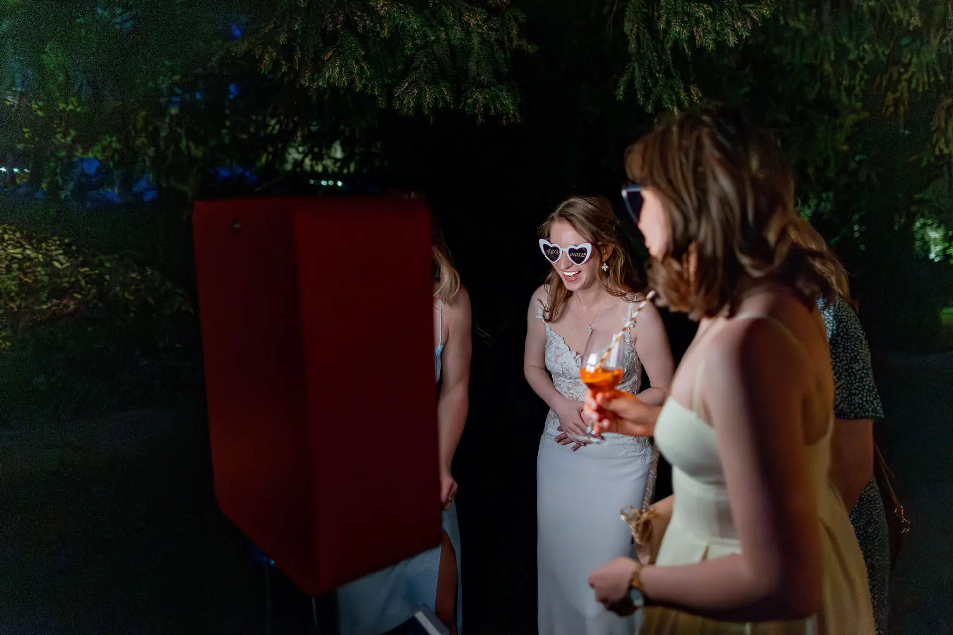 Braut mit herzförmiger Sonnenbrille und Gästen vor roter Fotobox bei Nacht