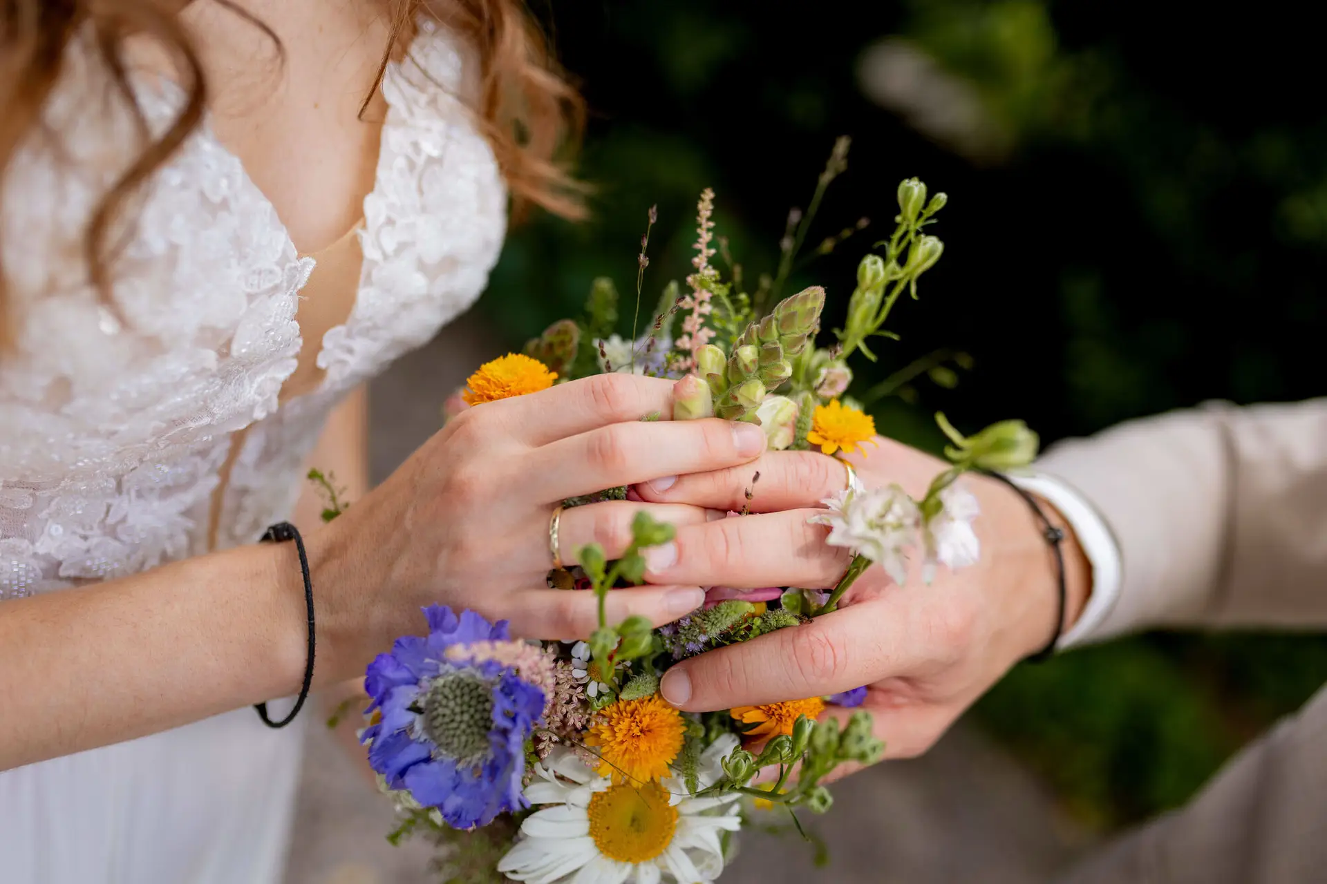 Hände des Brautpaares mit Eheringen auf dem bunten Wildblumenstrauß