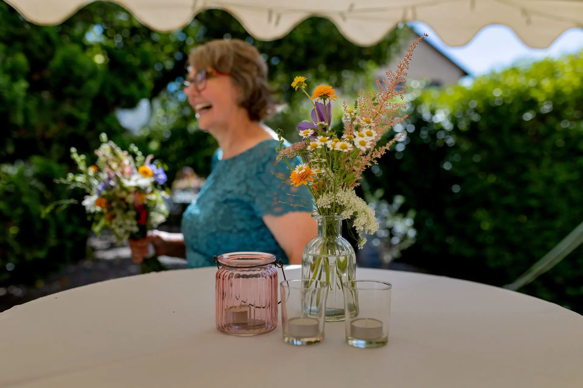 Stehtisch mit Blumenvase und Windlicht im Garten mit lachender Gästin