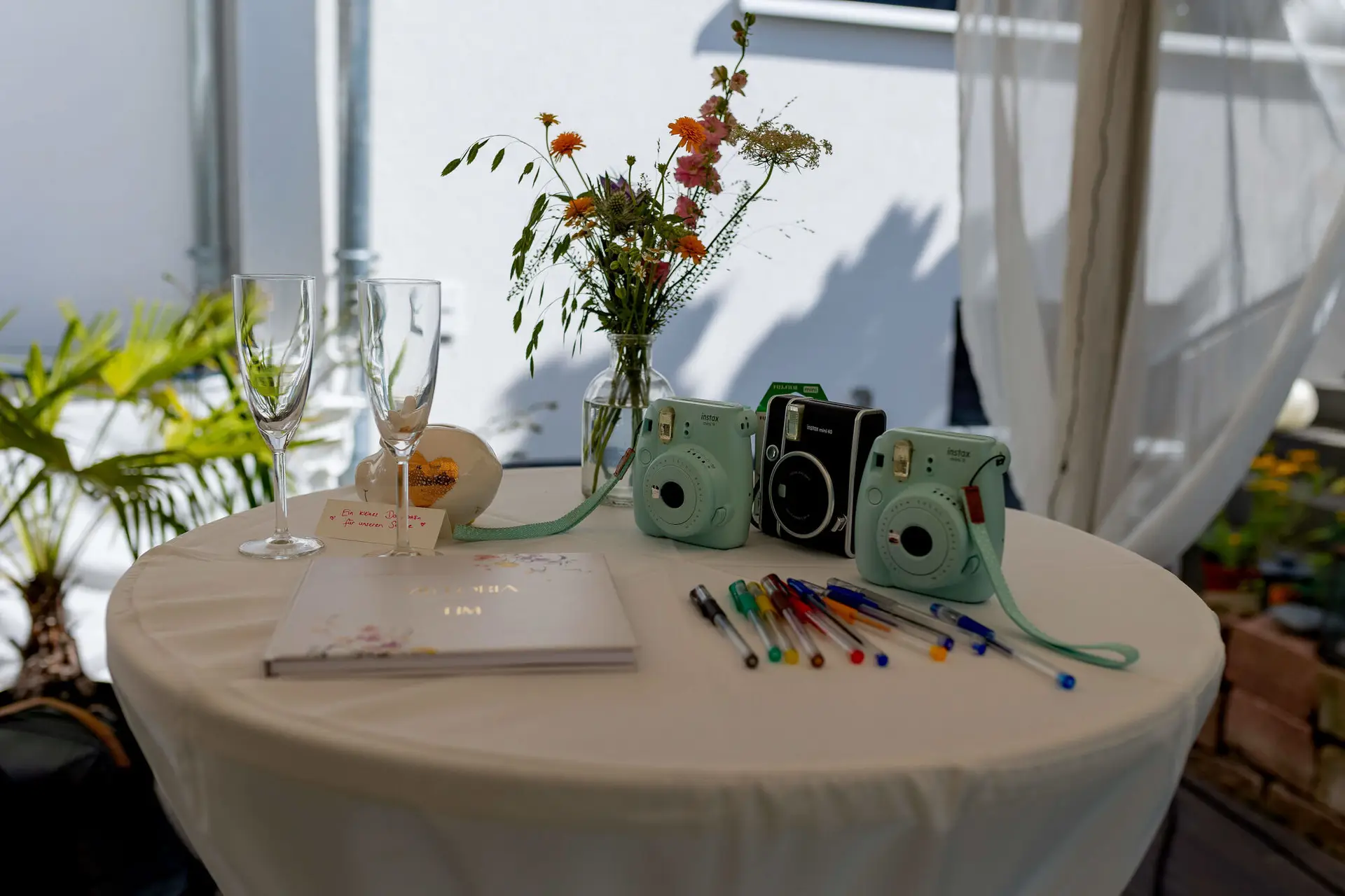 Gästebuch-Tisch mit Instax-Kameras, Blumenvase und Sektgläsern