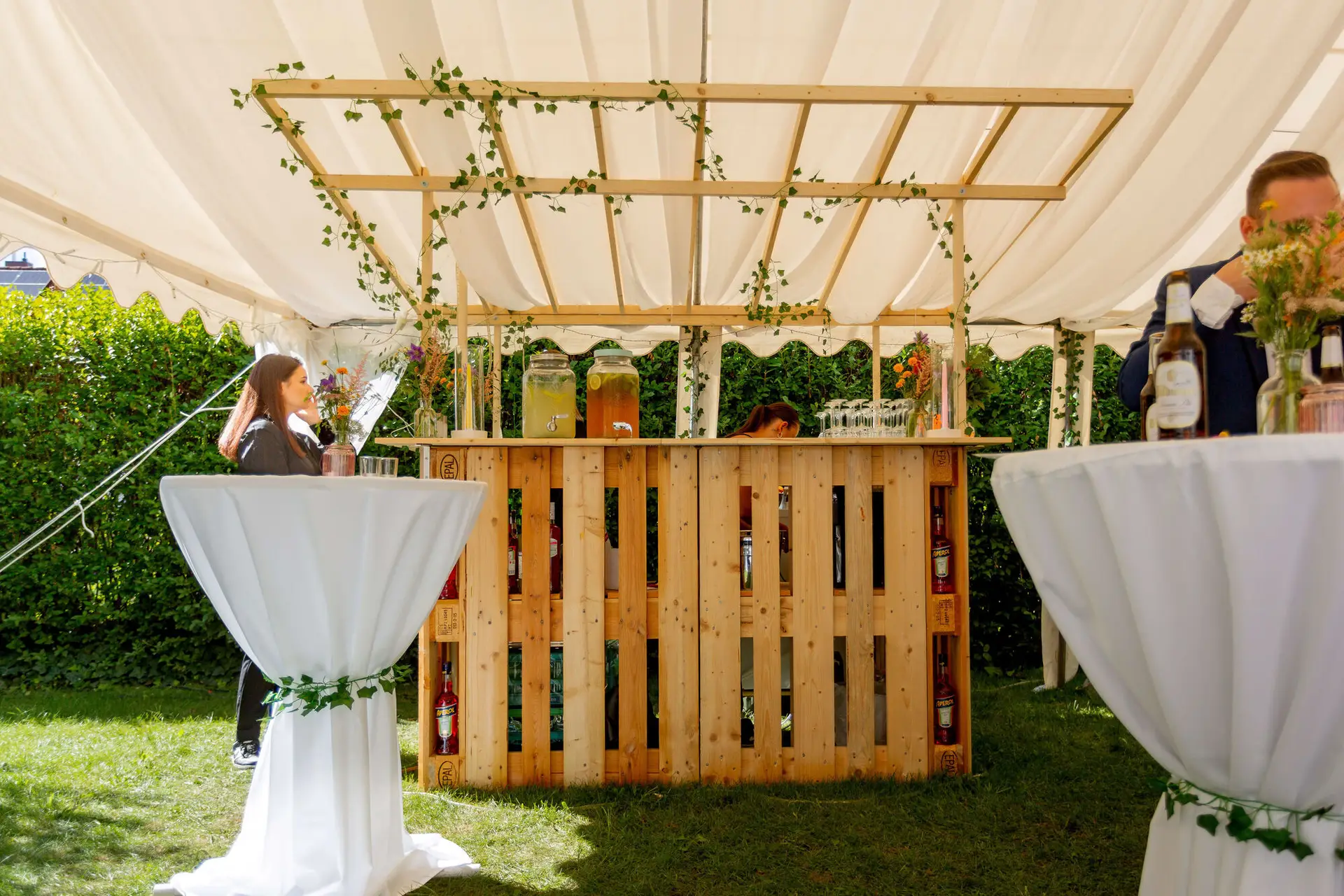 Selbstgebaute Bar aus Holzpaletten mit Efeuranken und Stehtischen unter dem Zelt