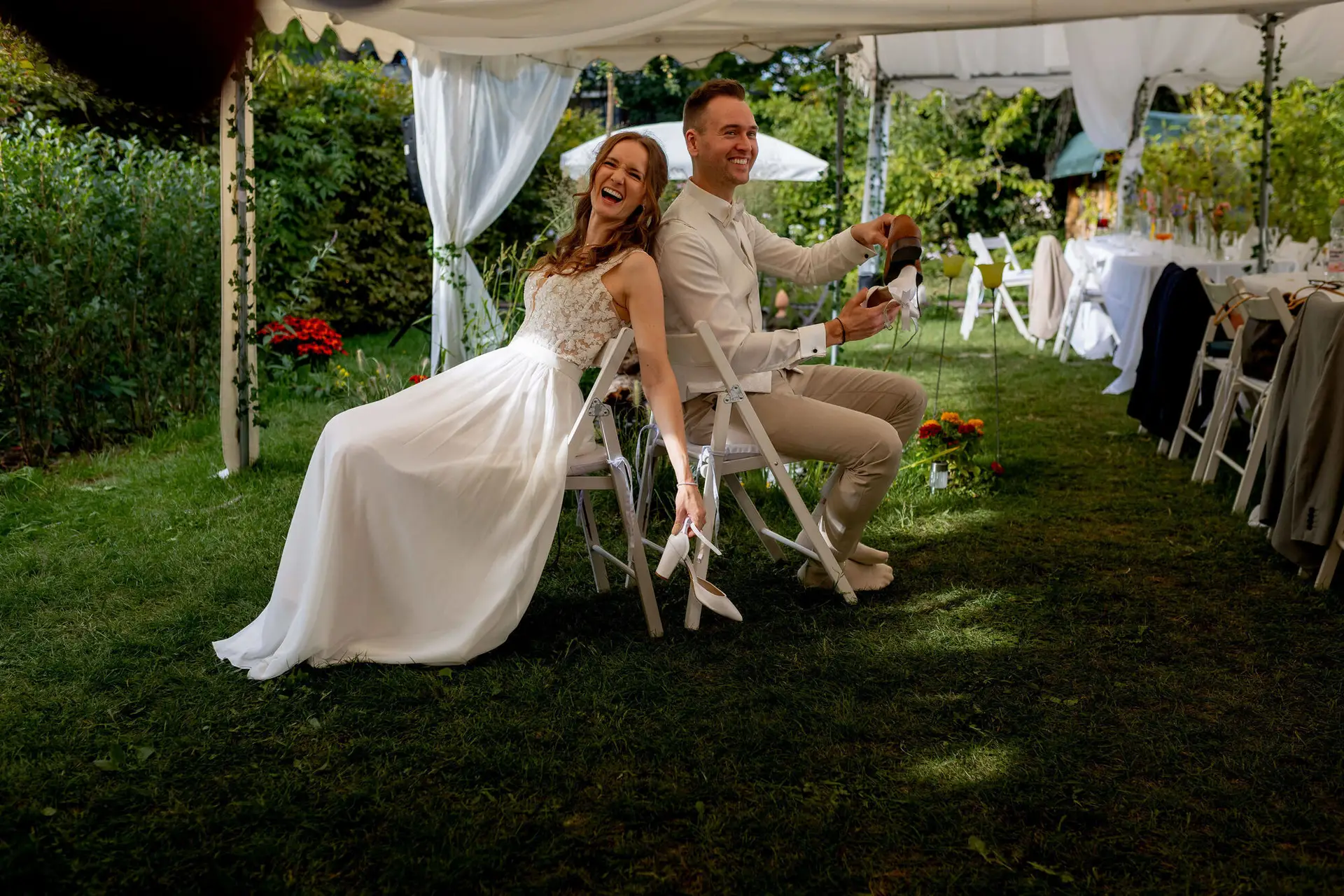 Brautpaar sitzt lachend beim Schuhspiel auf Klappstühlen im Garten