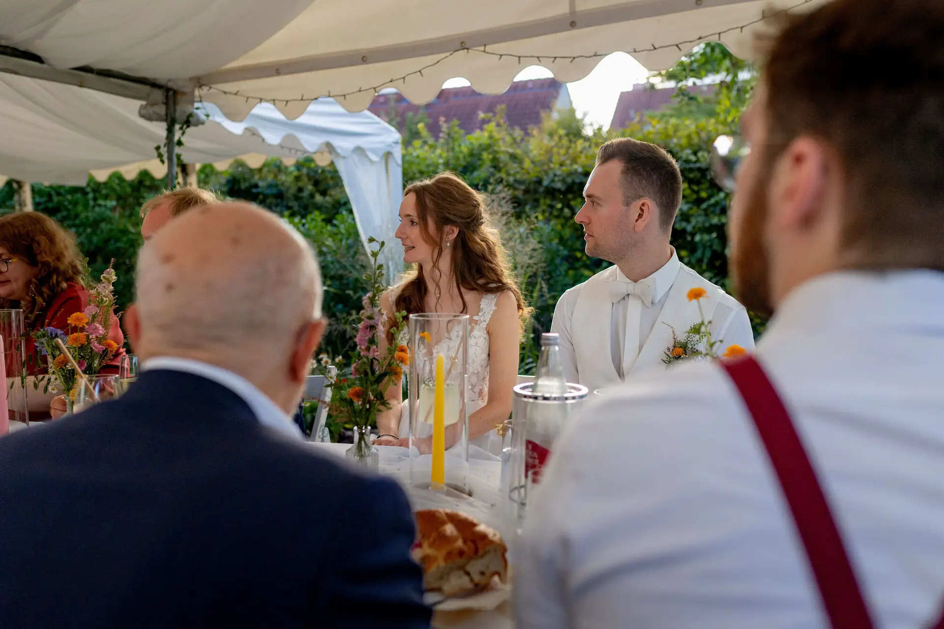 Brautpaar sitzt am gedeckten Tisch mit Gästen beim Abendessen im Gartenzelt