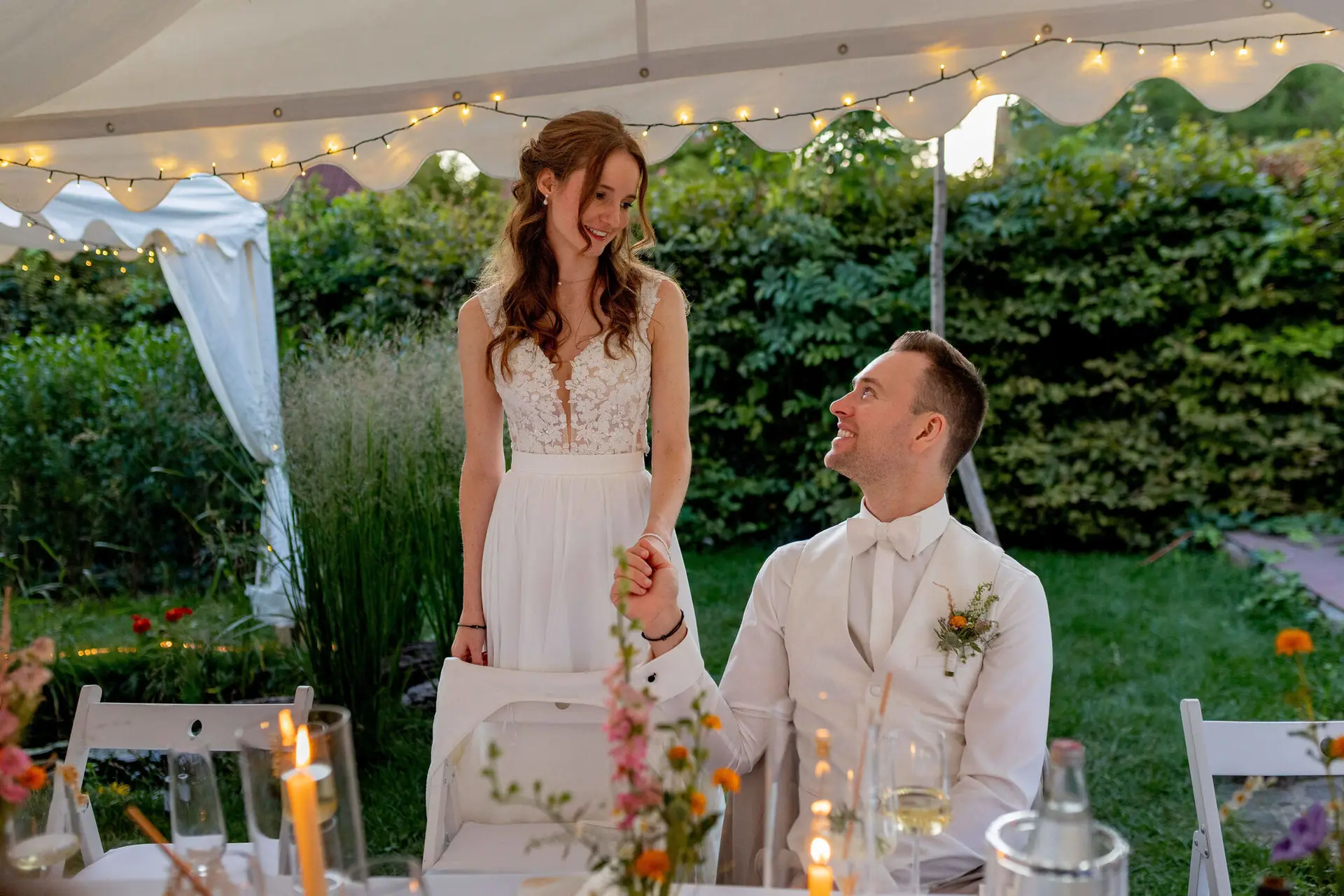 Braut steht lachend neben dem sitzenden Bräutigam im beleuchteten Zelt