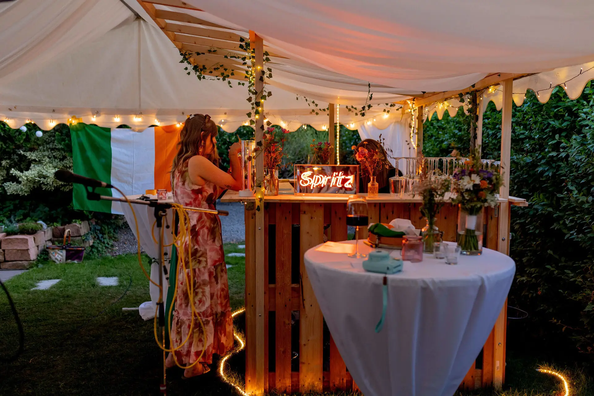 Beleuchtete Palettenbar mit Spritz-Neonschild und irischer Flagge am Abend
