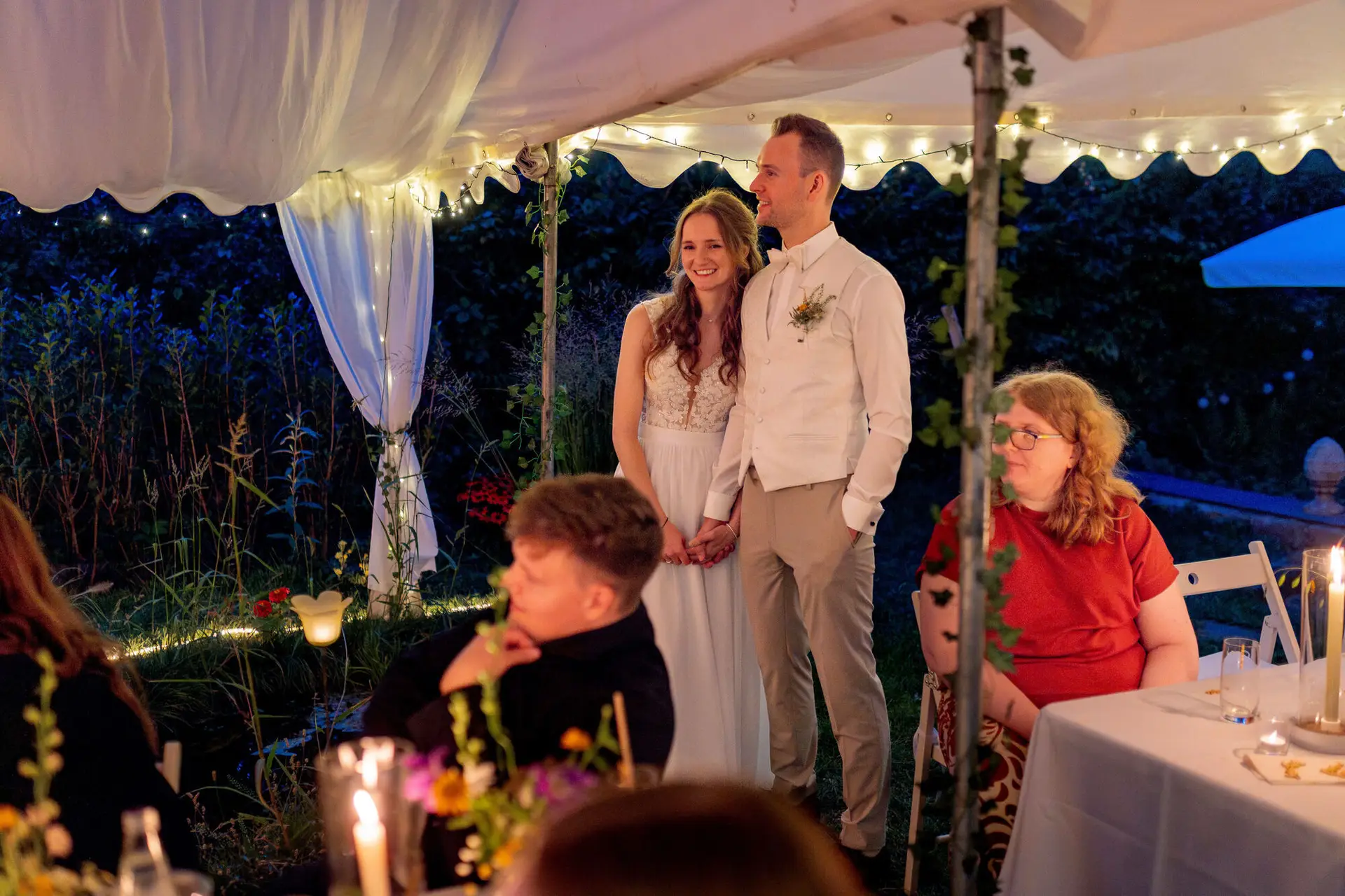 Brautpaar steht händchenhaltend vor den Gästen im beleuchteten Zelt am Abend