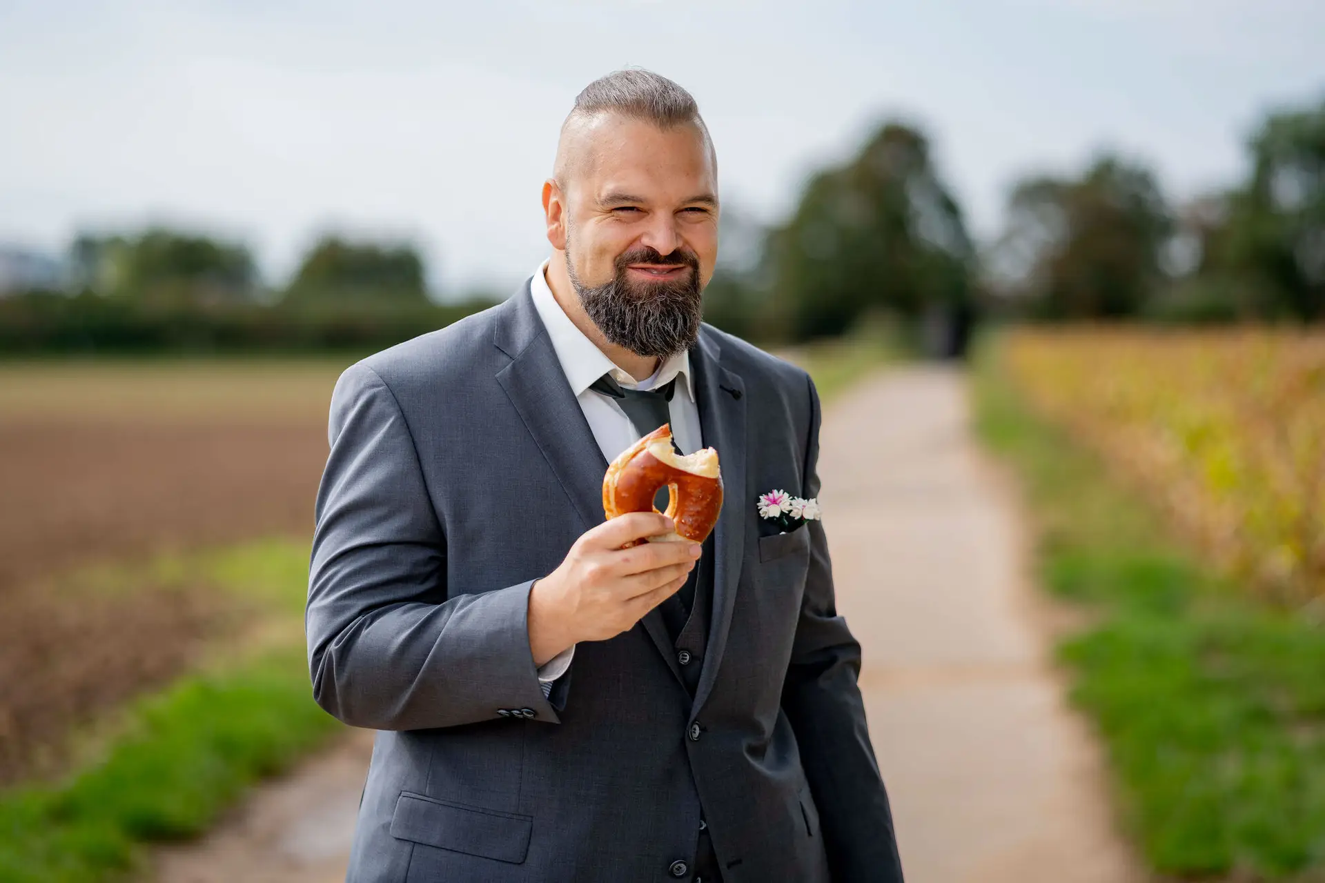Bräutigam im grauen Anzug isst eine Brezel auf einem Feldweg
