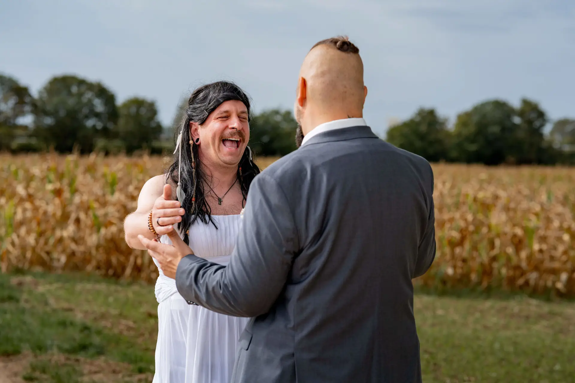Freund im weißen Kleid und Bräutigam lachen herzlich beim First-Look-Streich