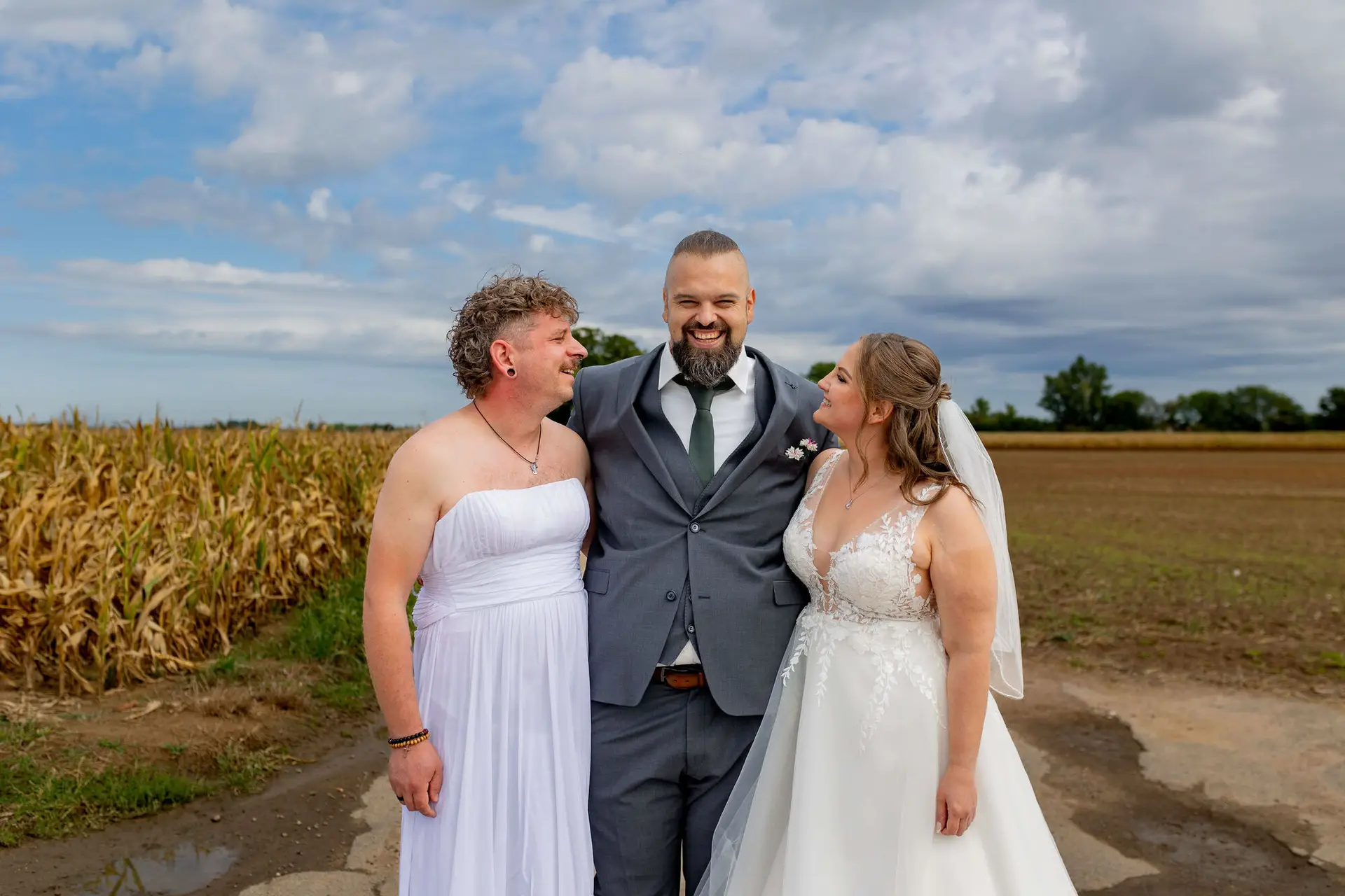 Bräutigam steht lachend zwischen Freund im weißen Kleid und Braut vor Kornfeld