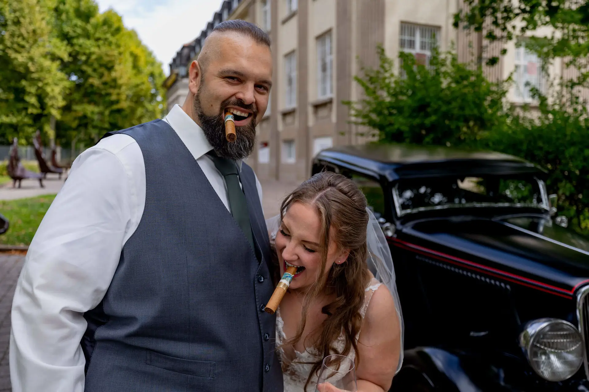 Brautpaar posiert lachend mit Zigarren vor dem Oldtimer
