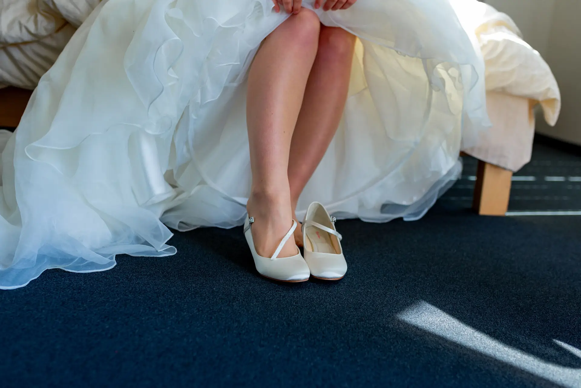 Braut sitzt auf dem Bett und zieht weiße Paillettenballerinas mit Schleife an