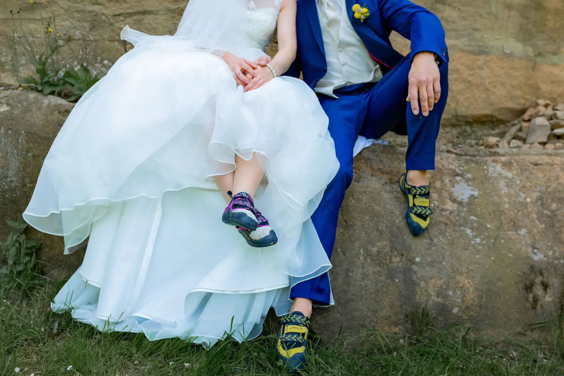 Braut und Bräutigam sitzen an einem Felsvorsprung, sie im weißen Kleid mit Wanderschuhen, er im blauen Anzug mit gelbem Blumenstrauß