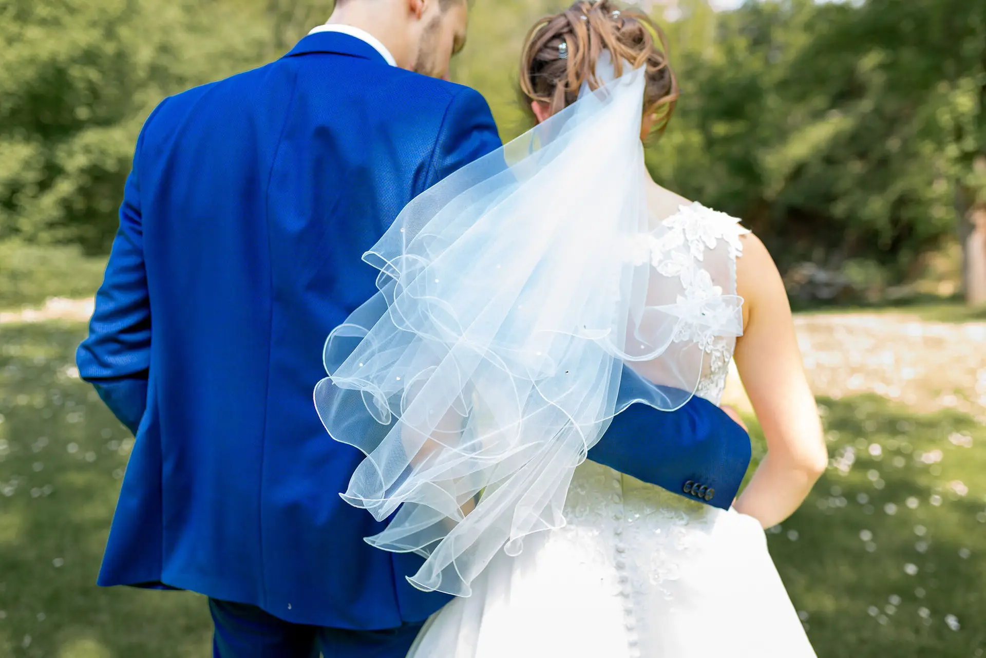 Brautpaar im Outdoor-Shooting, Bräutigam hält liebevoll den Schleier seiner Braut im Gegenlicht