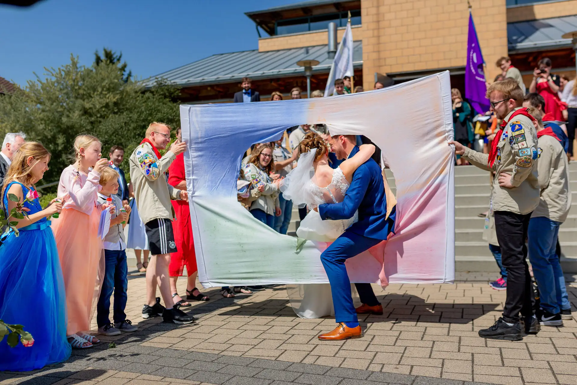 Brautpaar küsst sich leidenschaftlich unter buntem Stoffbanner vor blauem Himmel, Gäste jubeln rundherum